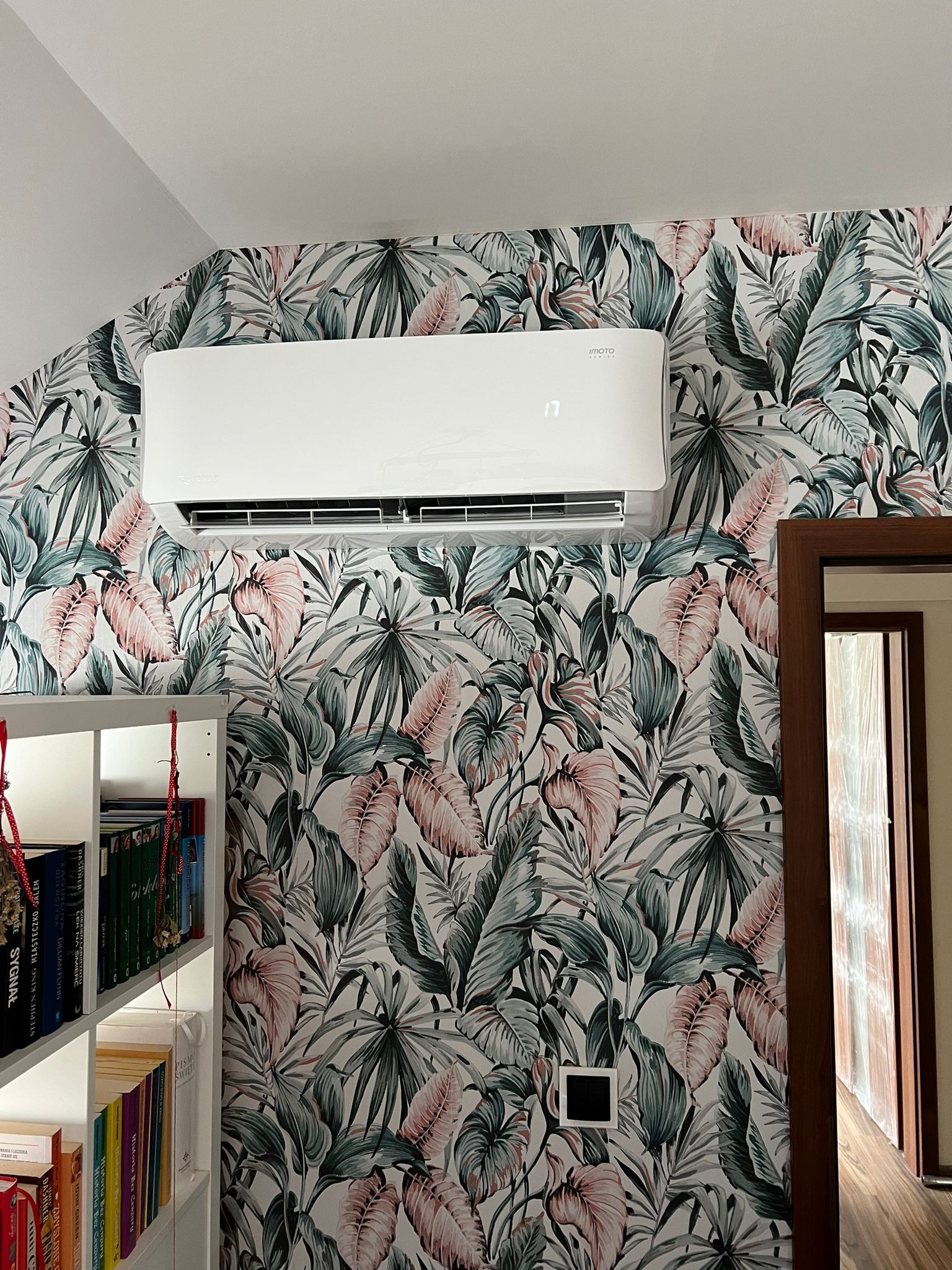 Biały klimatyzator na ścianie w pokoju z tropikalną tapetą, obok regał z książkami i framuga drzwi.