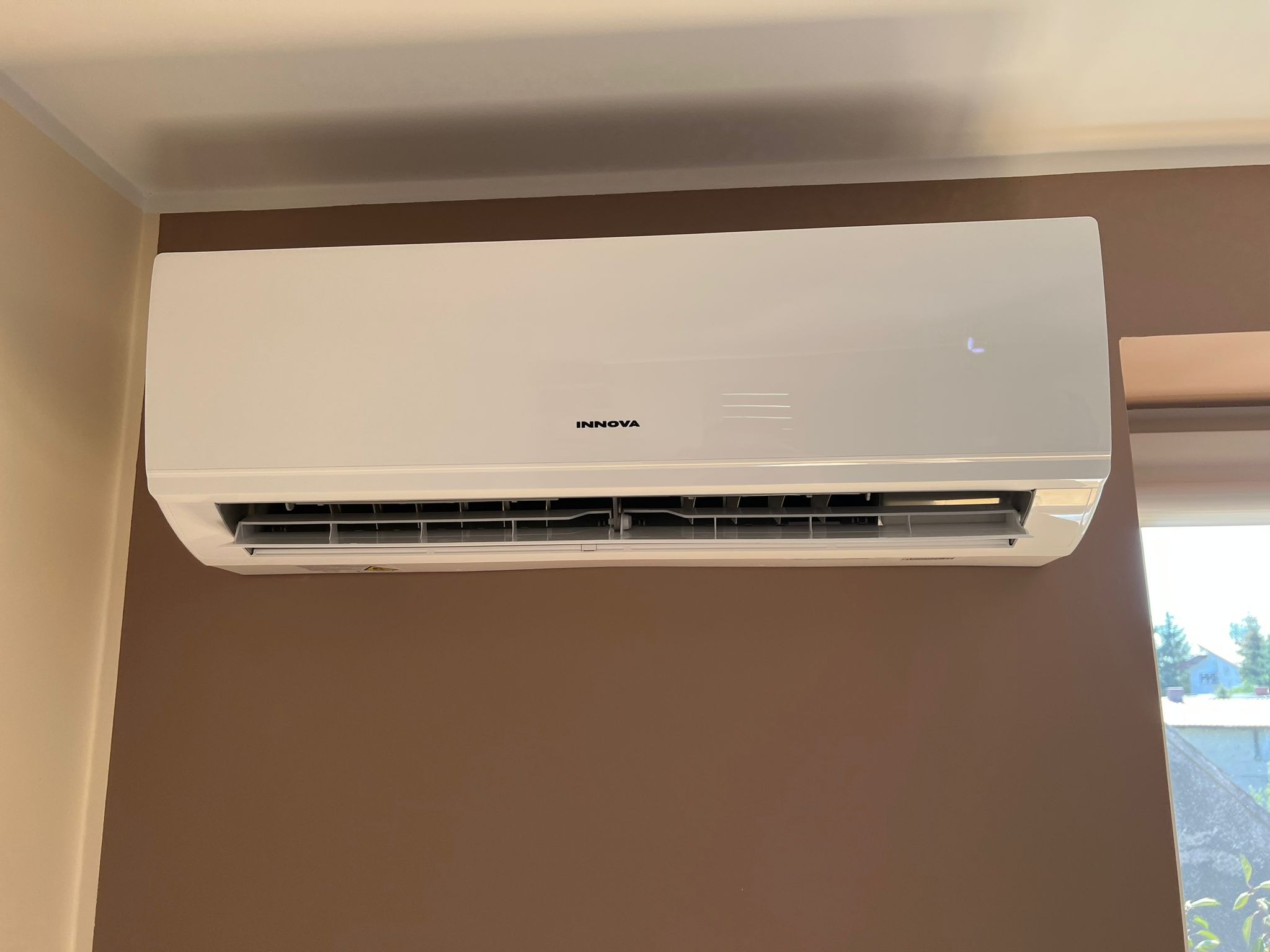 Biały klimatyzator marki Innova zamontowany na brązowej ścianie w pomieszczeniu, widok z dołu, z widocznym fragmentem okna i zielenią na zewnątrz.