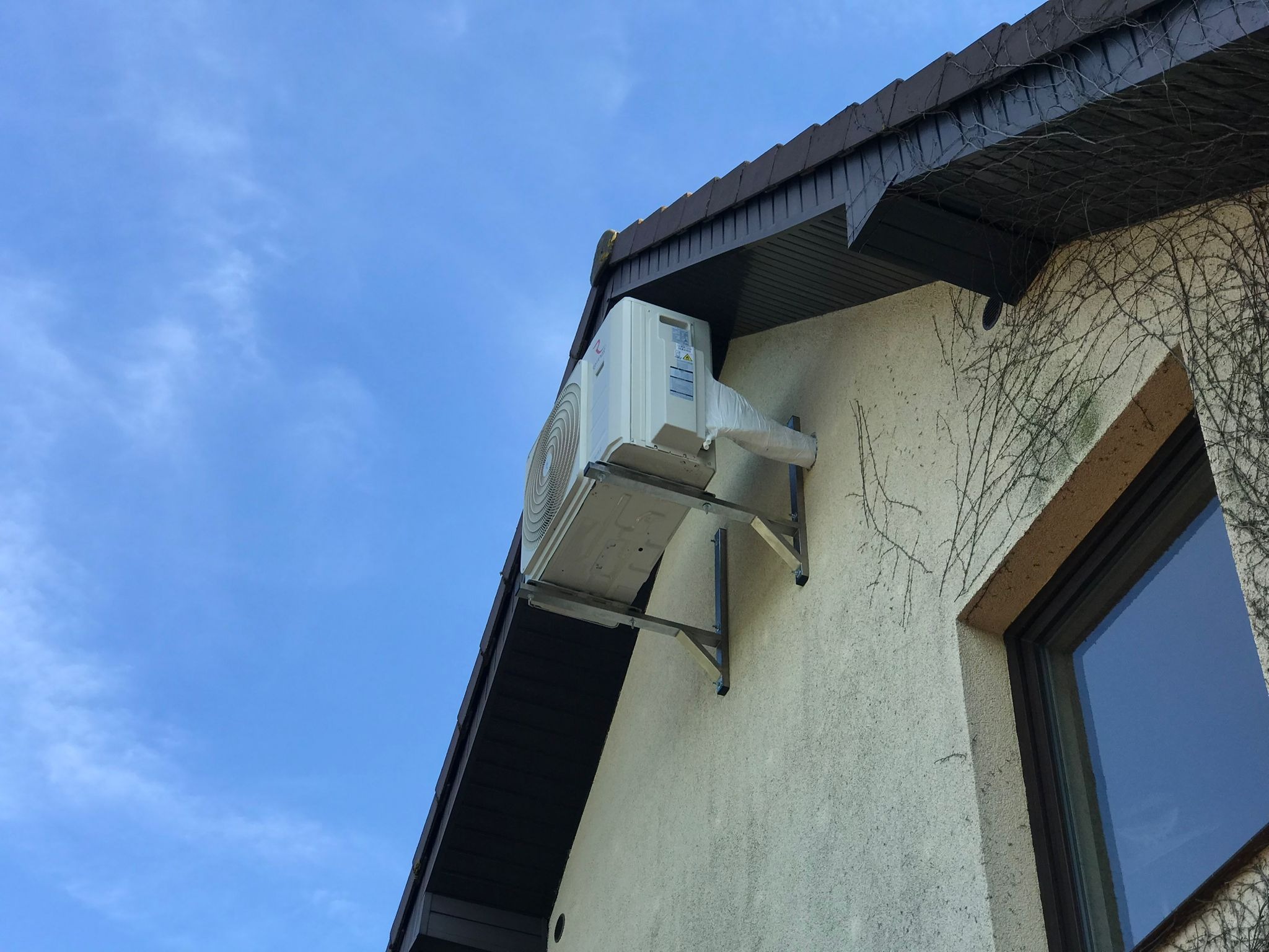 Zewnętrzna jednostka klimatyzatora zamontowana na metalowym wsporniku, częściowo owinięta folią ochronną, widoczna na tle ściany budynku z oknem i błękitnego nieba.