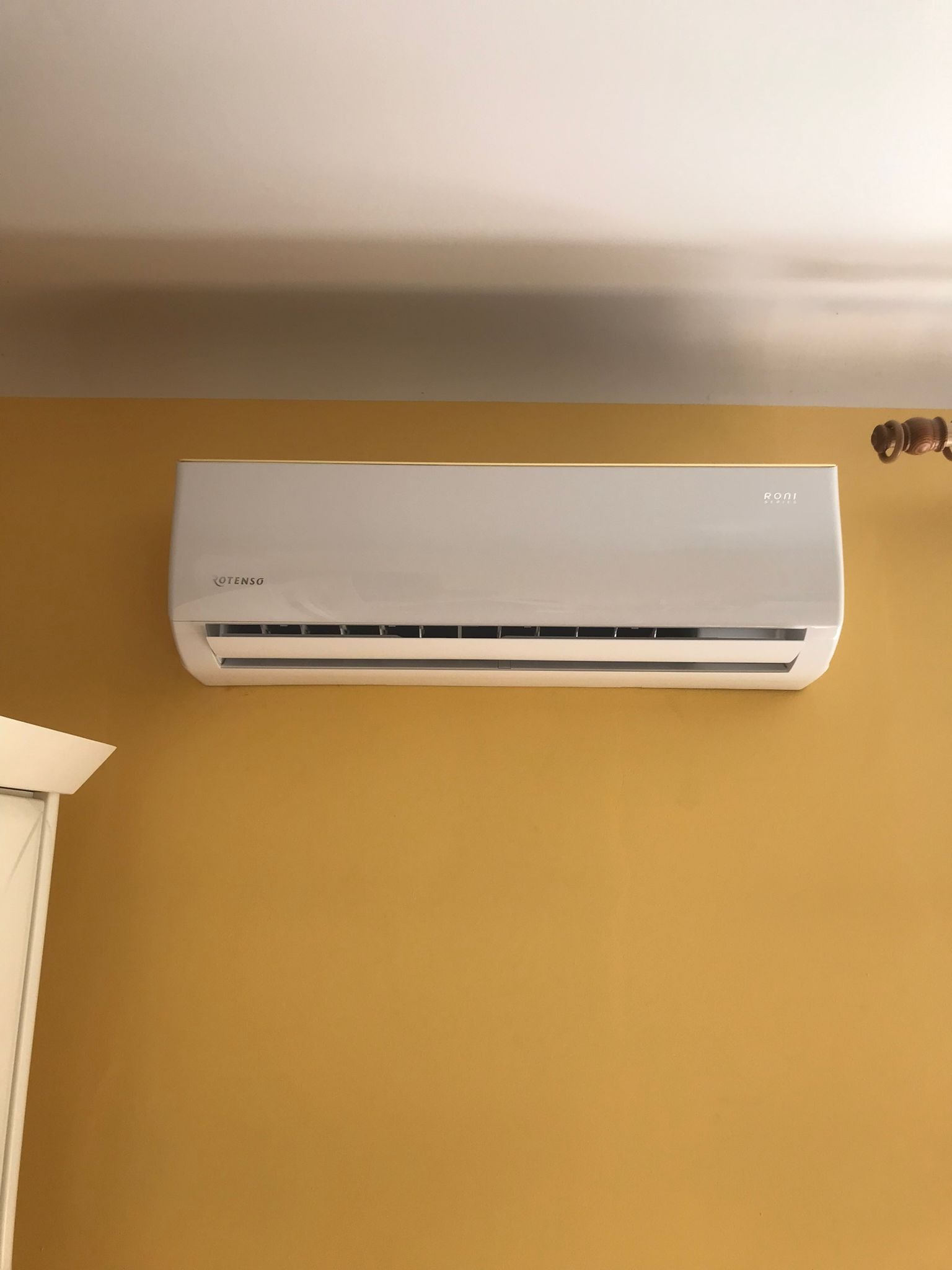 Biała jednostka klimatyzacyjna marki Rotenso Roni zamontowana na musztardowej ścianie w pomieszczeniu mieszkalnym, widoczny fragment listwy przypodłogowej i sufitu.