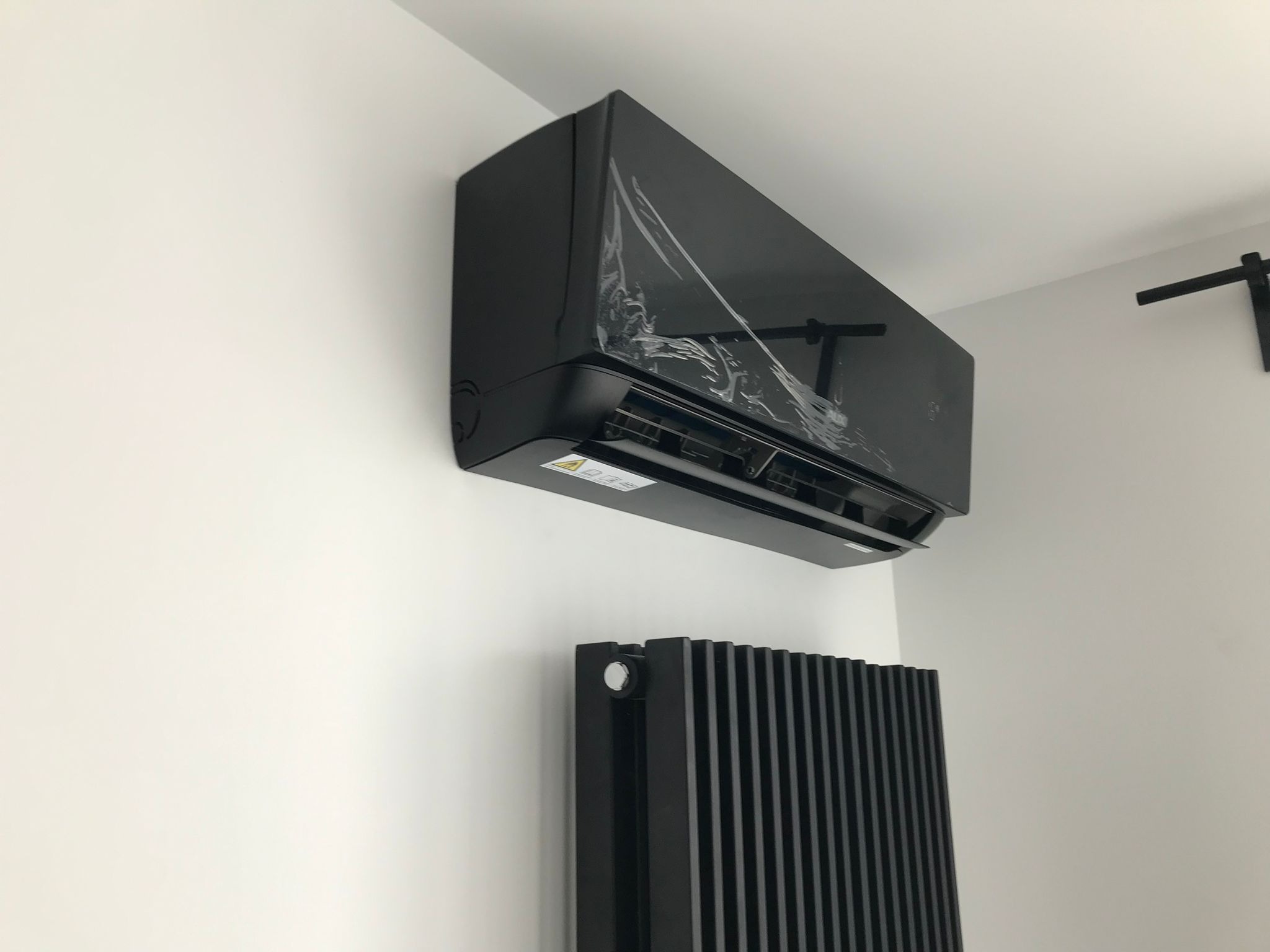 Czarny klimatyzator ścienny, jeszcze z folią ochronną, zamontowany nad czarnym grzejnikiem żeberkowym na białej ścianie, widoczny fragment drążka do podciągania.
