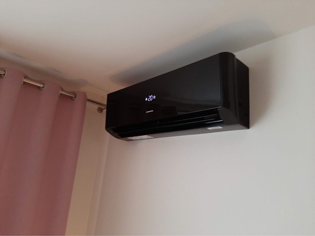 Czarny klimatyzator ścienny włączony, wyświetla temperaturę 20 stopni, widoczny fragment różowej zasłony.
