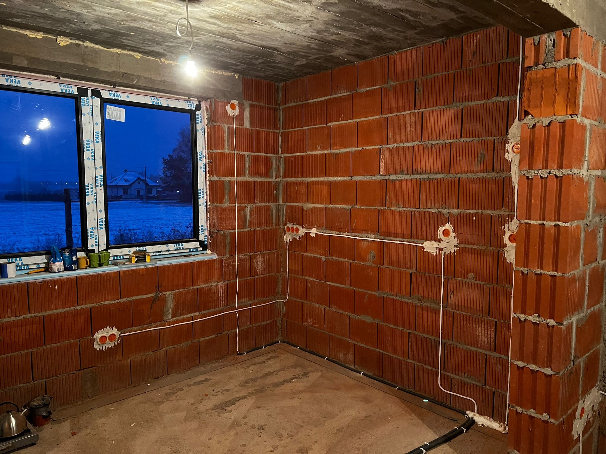 Surowy stan wnętrza z pomarańczowej cegły dziurawki. Widoczne okno z widokiem na ośnieżony krajobraz. Na ścianach i podłodze poprowadzone kable elektryczne w peszlu i puszki pod gniazdka, w tle...