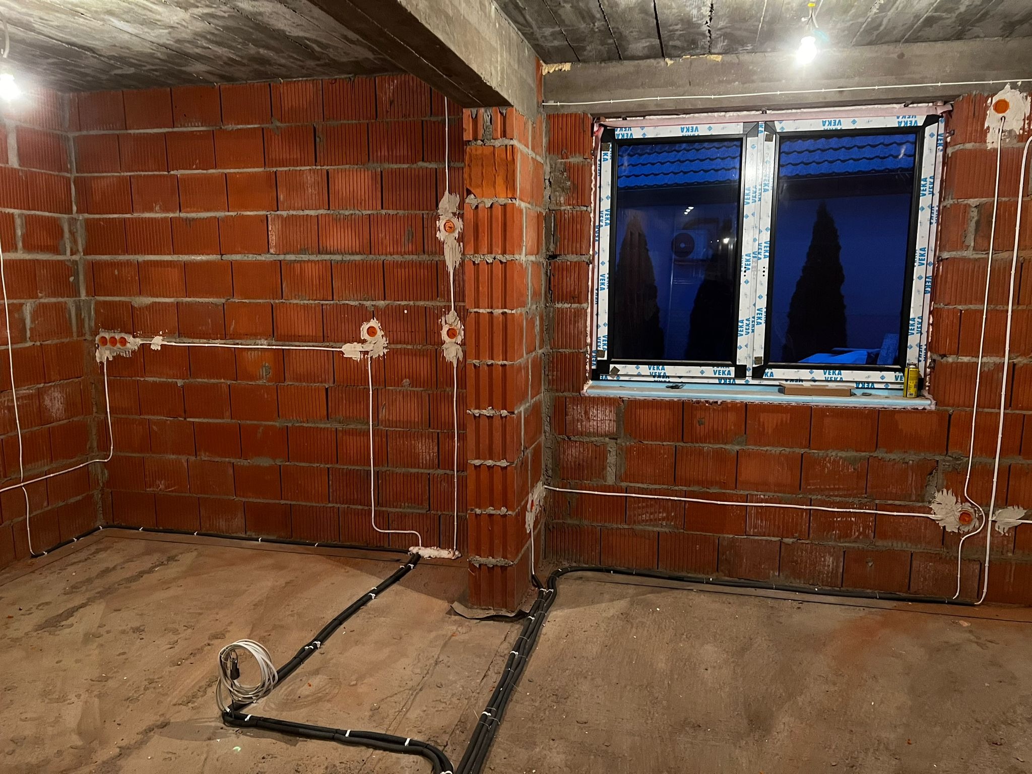 Surowy stan wnętrza budynku z widoczną instalacją elektryczną: białe przewody podtynkowe i czarne rury karbowane prowadzone po podłodze, czerwona cegła dziurawka na ścianach, okno PCV z widokiem...