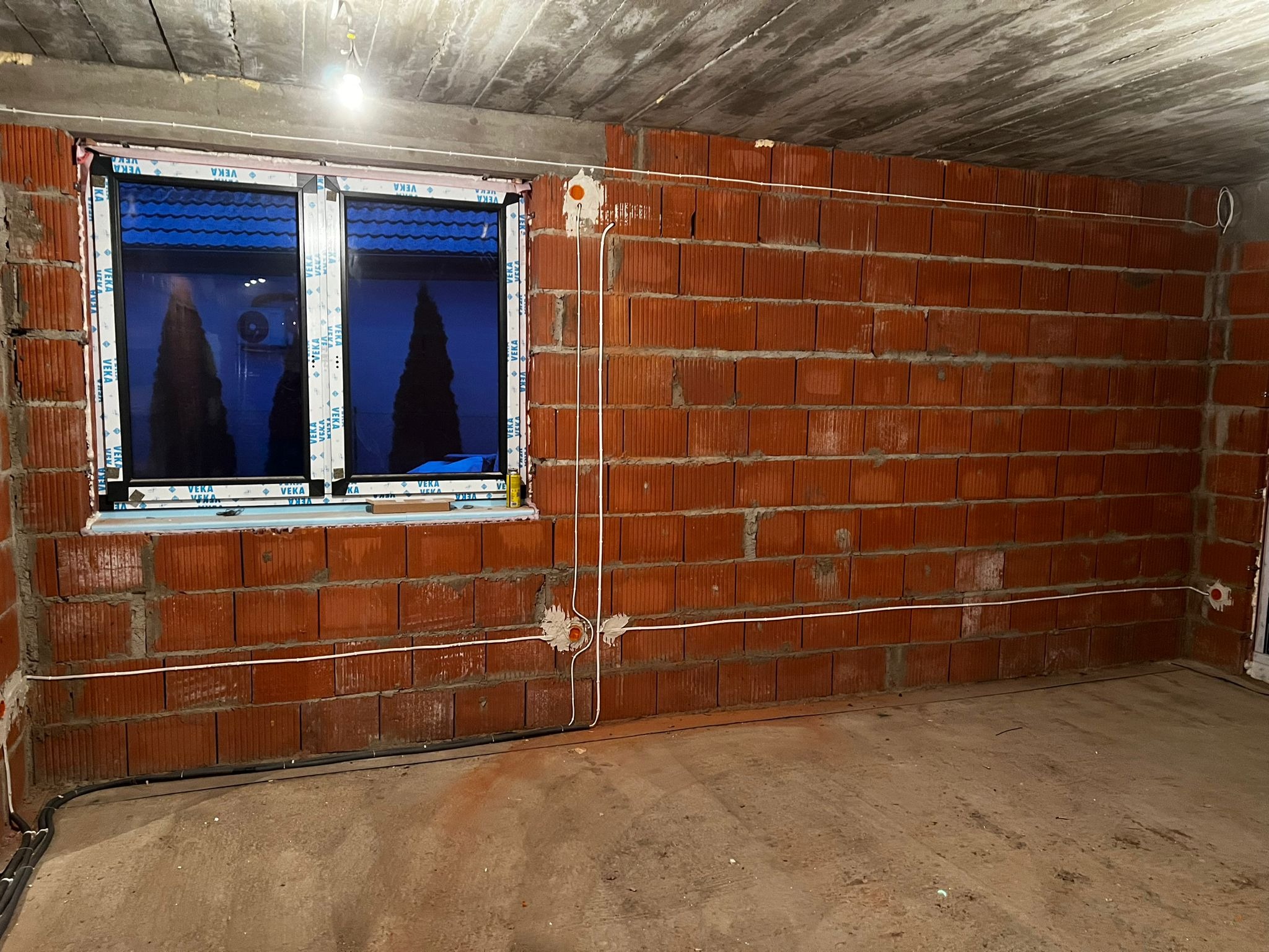 Wnętrze w trakcie remontu: surowy betonowy sufit, ściany z czerwonej cegły dziurawki z widocznymi przewodami elektrycznymi w białej izolacji, okno z widocznym klimatyzatorem na zewnątrz. Widoczne...