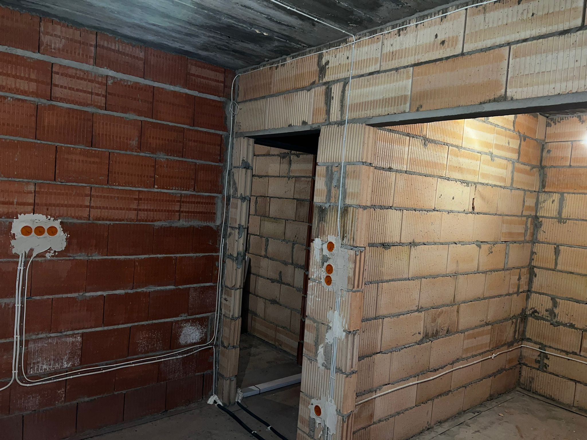 Surowy stan wnętrza w trakcie prac elektrycznych: widoczne poprowadzone przewody w ścianach z cegły dziurawki, puszki elektryczne z otworami, betonowy sufit.