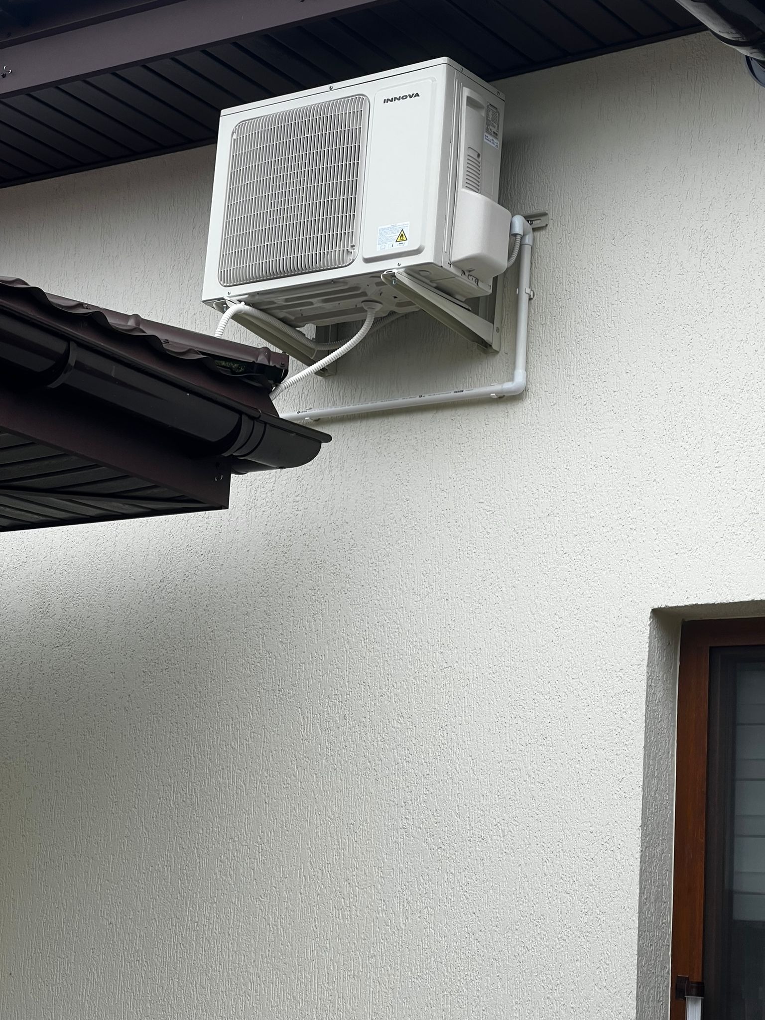 Biała jednostka klimatyzacji Innova zamontowana na jasnej, teksturowanej ścianie zewnętrznej domu, widoczna rynna dachowa i fragment okna.