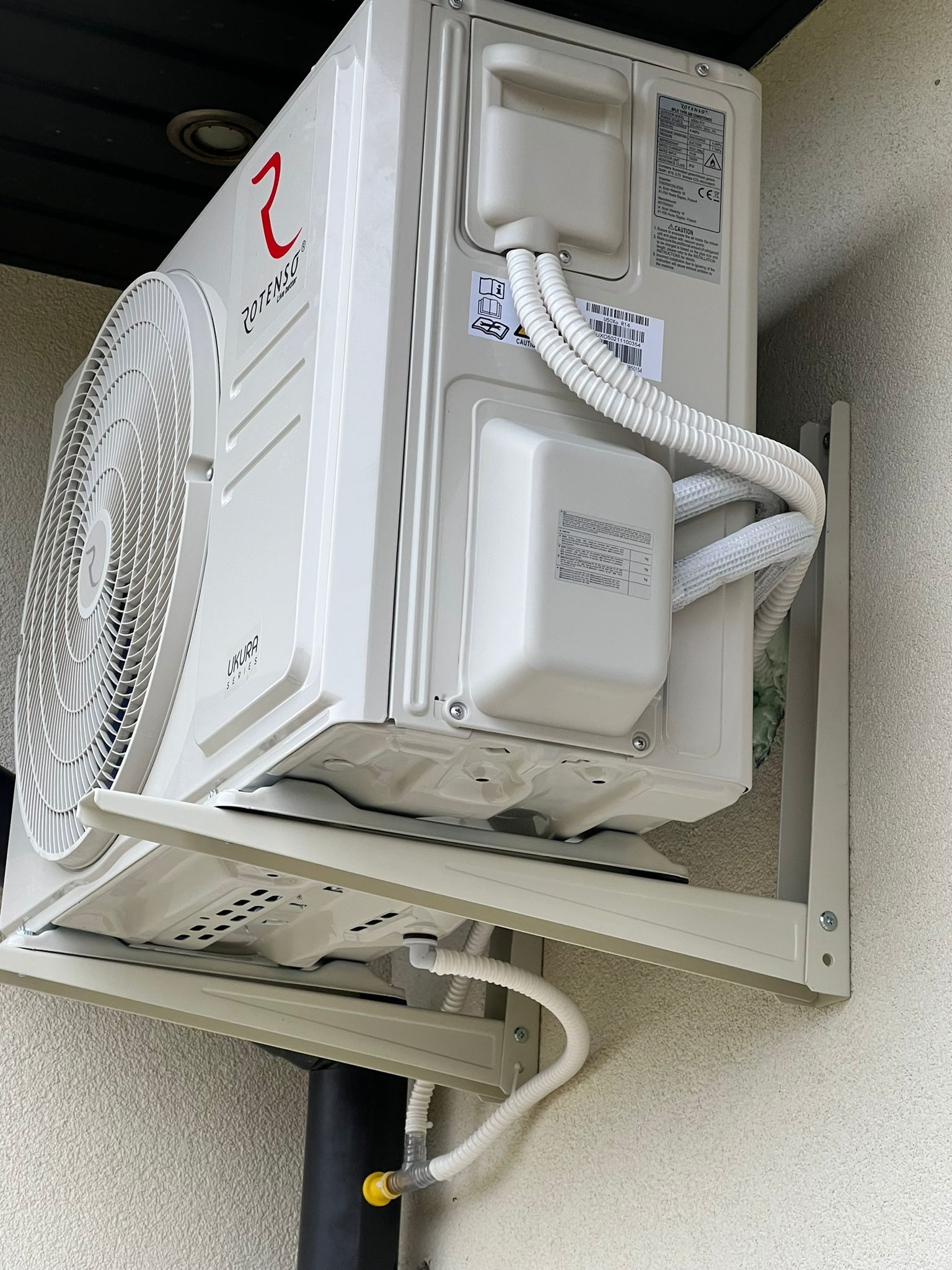 Zewnętrzna jednostka klimatyzacji Rotenso Ukura Series, zamontowana na ścianie budynku, widoczne białe rury odprowadzające skropliny i przewody w peszlach, wspornik montażowy w kolorze obudowy.