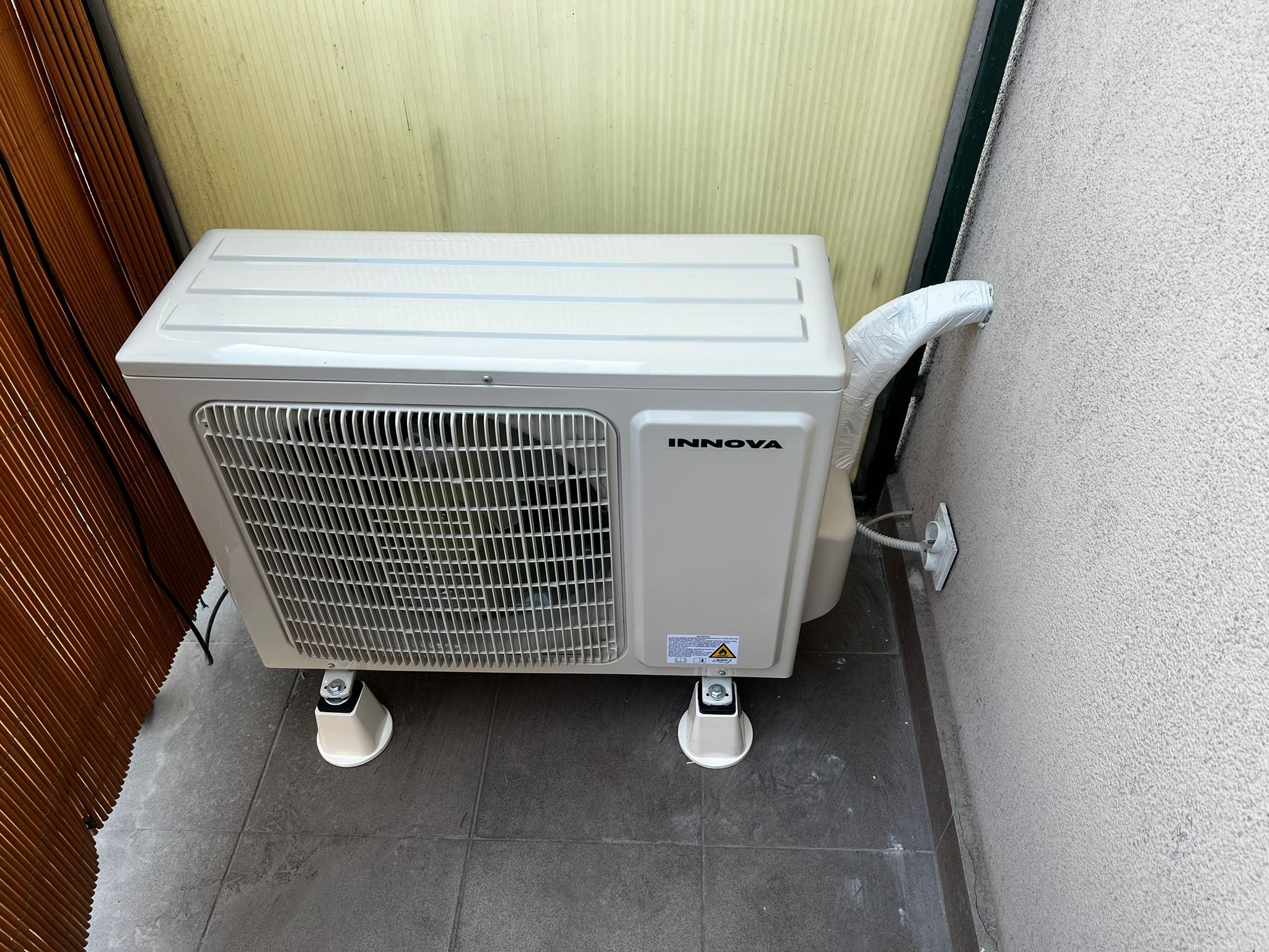 Zewnętrzna jednostka klimatyzacji Innova, zamontowana na czterech białych wspornikach na szarych płytkach, z widocznym przewodem odprowadzającym skropliny owiniętym folią, z tyłu widoczna beżowa...