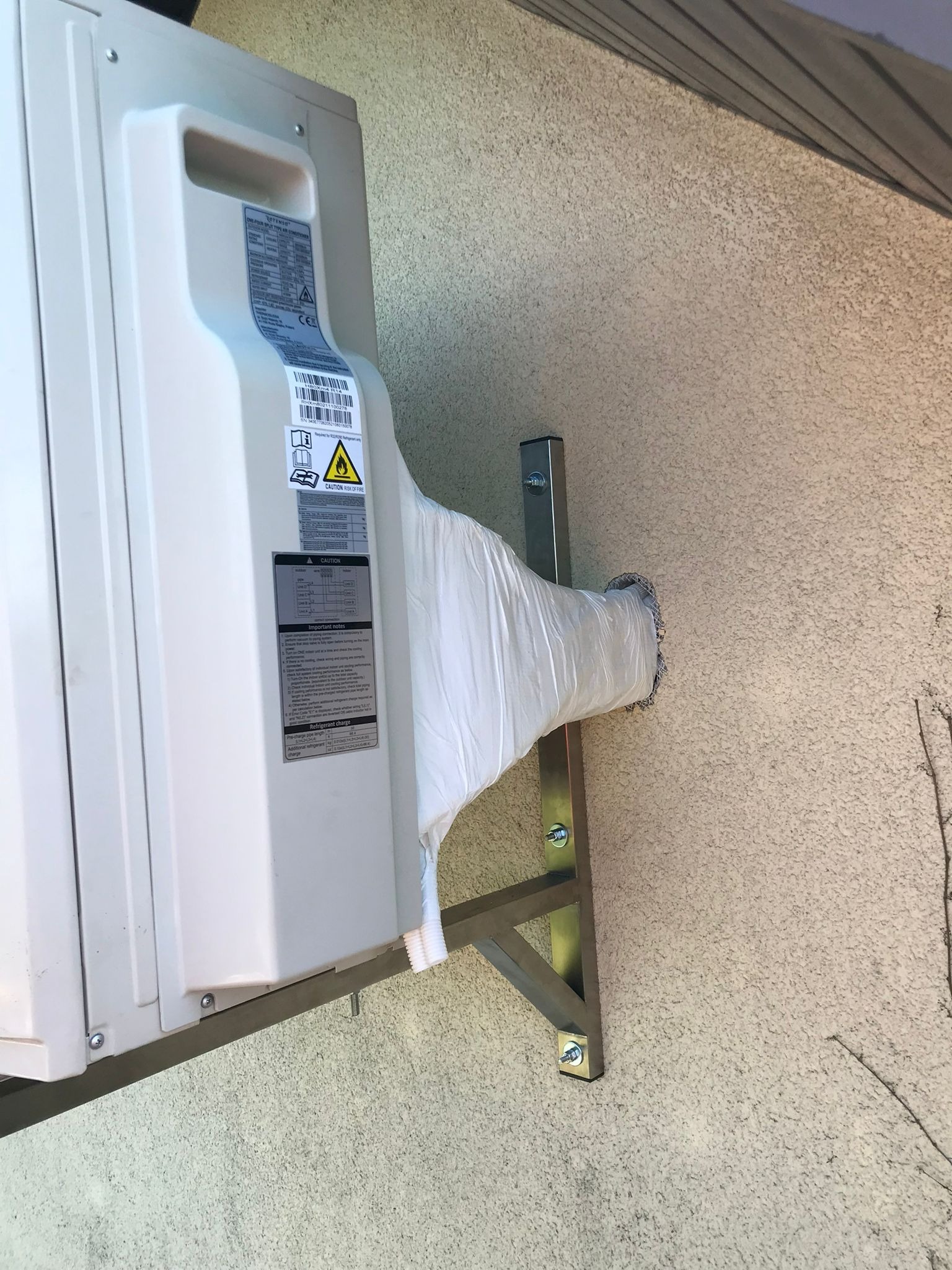 Zewnętrzna jednostka klimatyzatora zamontowana na metalowym wsporniku na elewacji budynku, z widocznym przewodem odprowadzającym skropliny owiniętym folią ochronną.