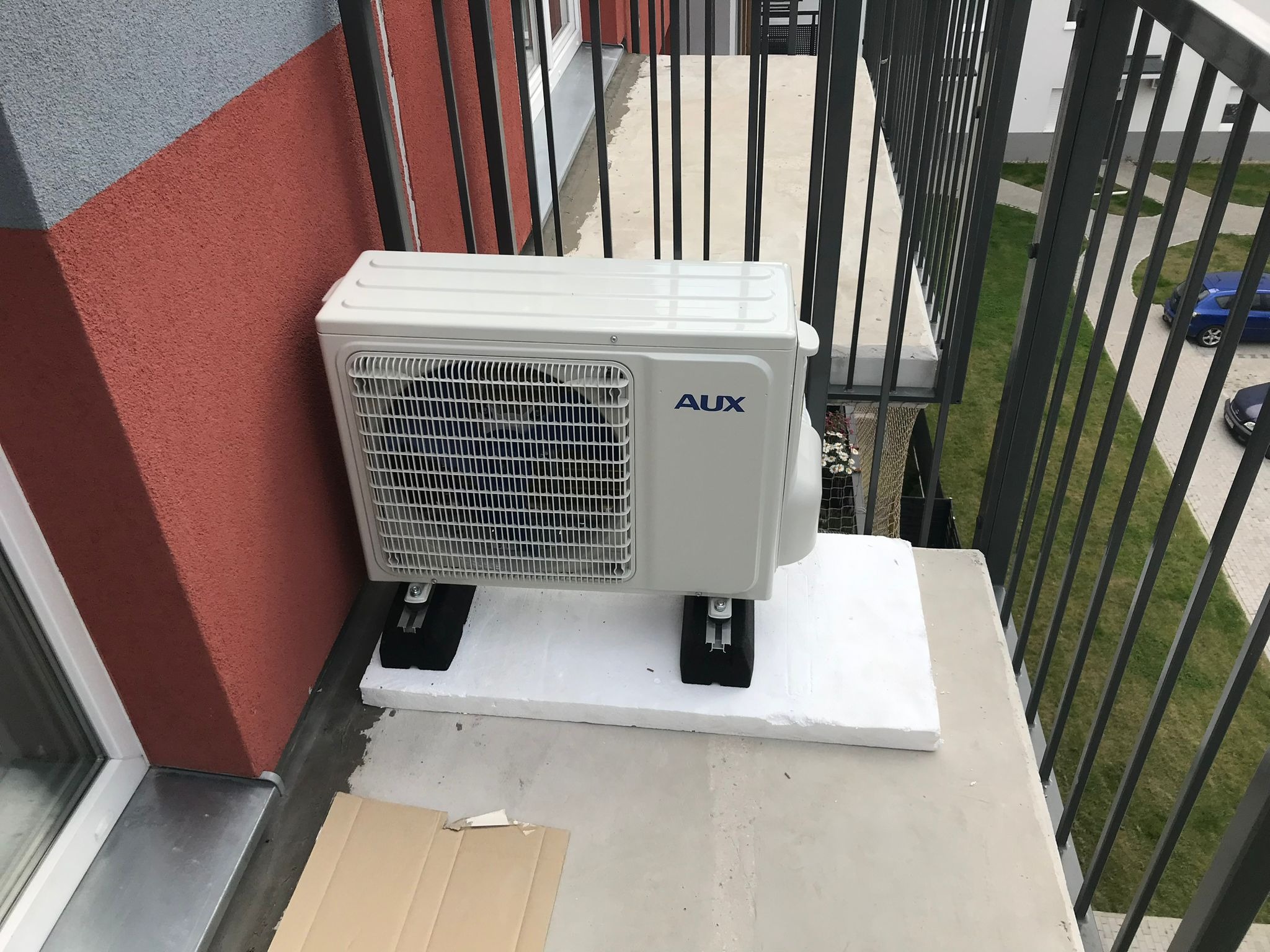 Biała jednostka zewnętrzna klimatyzacji AUX zamontowana na balkonie bloku mieszkalnego, umieszczona na białej styropianowej podkładce z czarnymi gumowymi nóżkami antywibracyjnymi, widoczne elementy...
