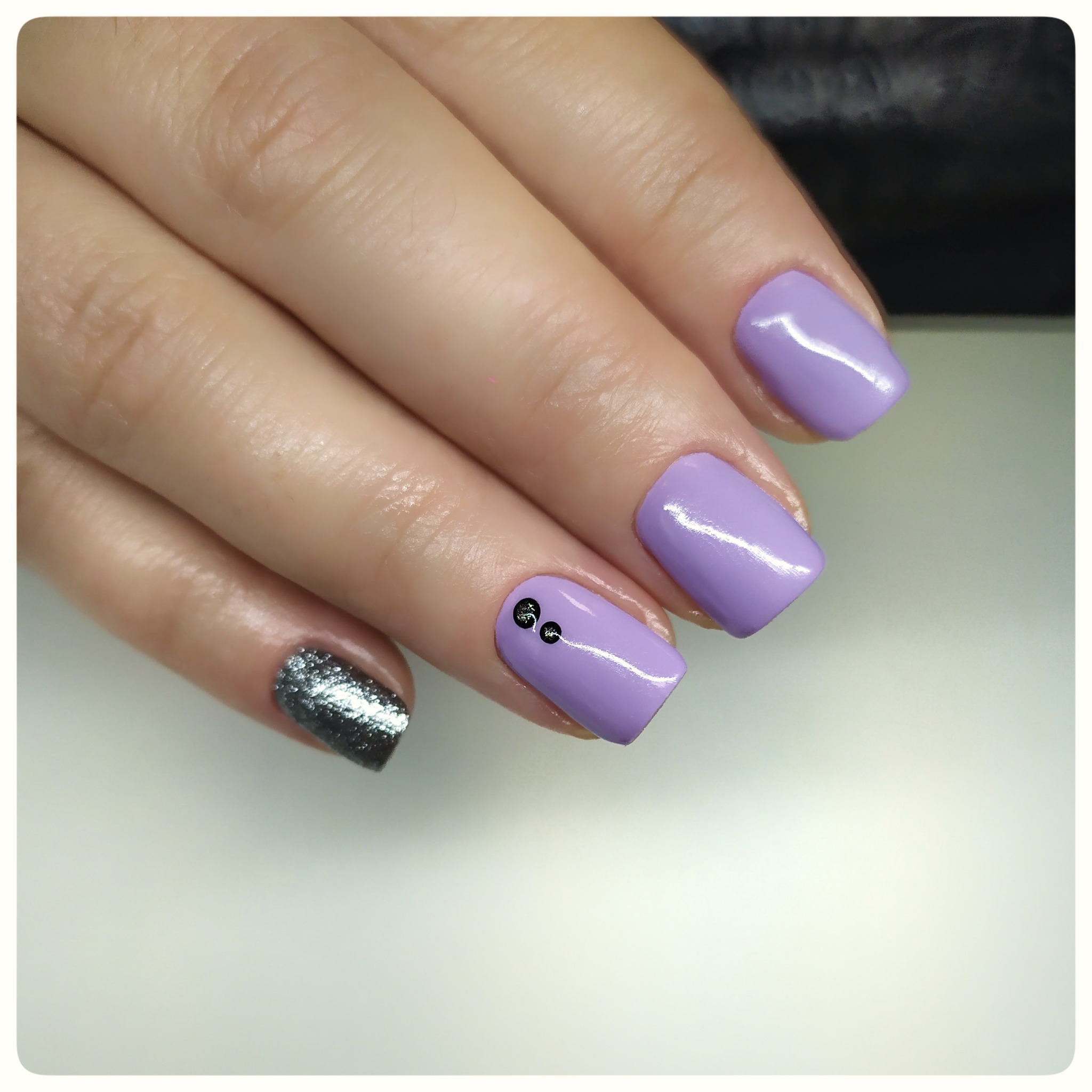 Dłoń z pomalowanymi paznokciami: jeden srebrny brokatowy, pozostałe pastelowo fioletowe z drobnymi czarnymi zdobieniami.