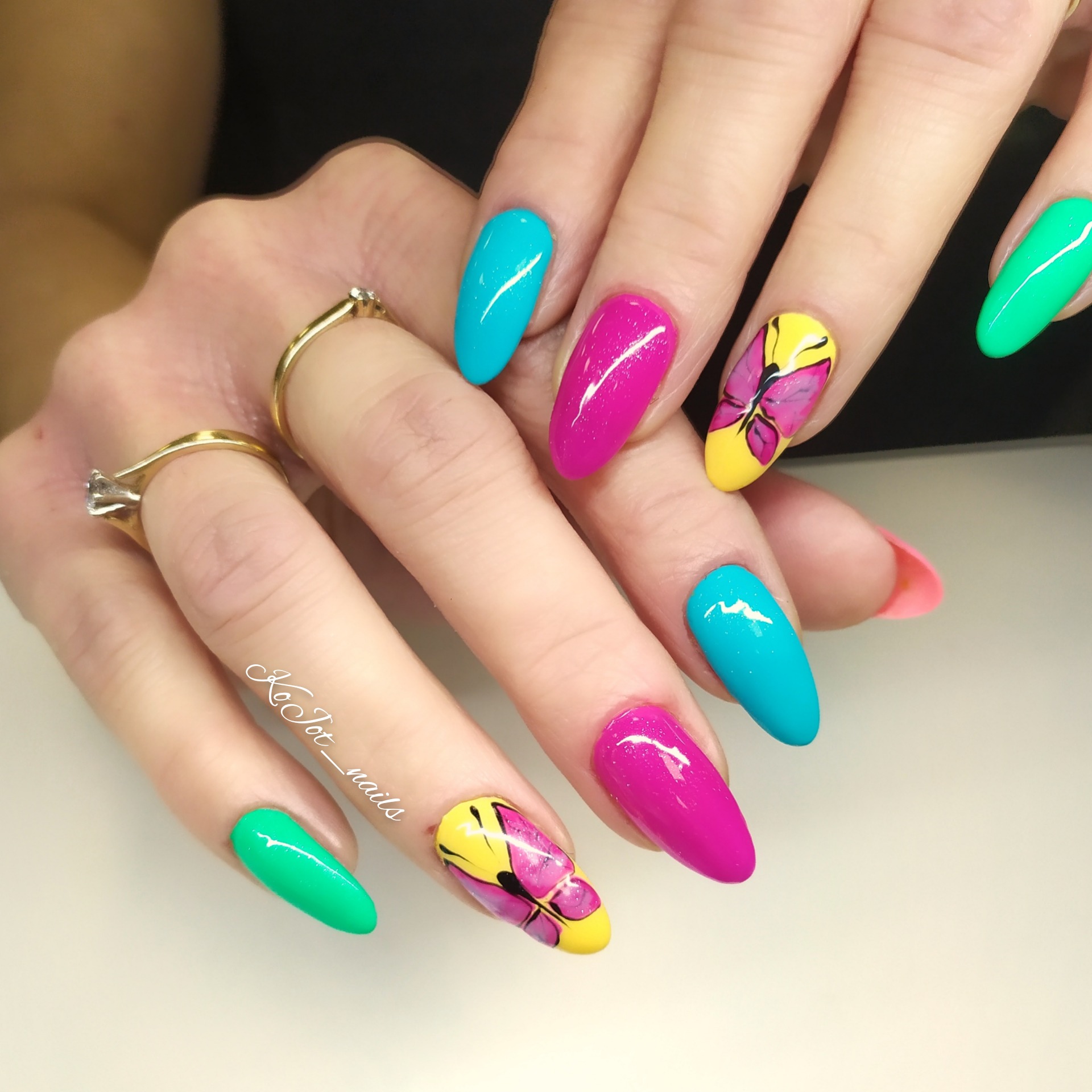 Paznokcie w kolorach: turkus, fuksja i limonka, zdobione motywem motyla na żółtym tle, dłonie ozdobione złotymi pierścionkami