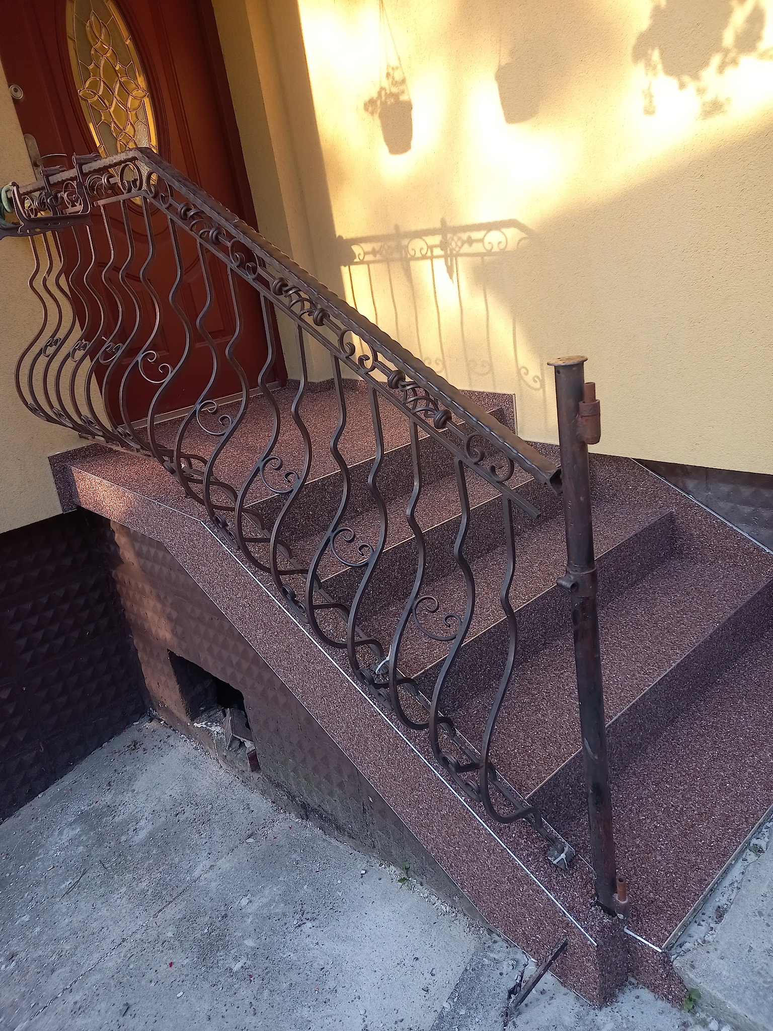 Zewnętrzne schody wejściowe z brązowego lastryko z ozdobną, kutą balustradą, prowadzące do drzwi wejściowych. Na ścianie widoczne cienie wiszących doniczek.