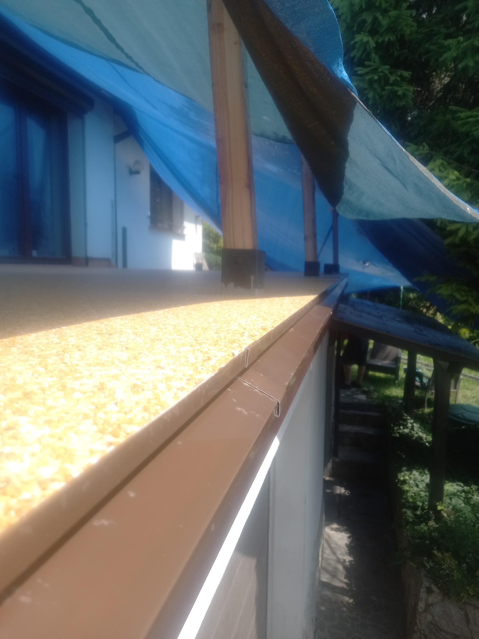 Wykończona powierzchnia balkonu z brązowym obramowaniem, zabezpieczona niebieską plandeką wspartą na drewnianych słupkach, widok z boku.