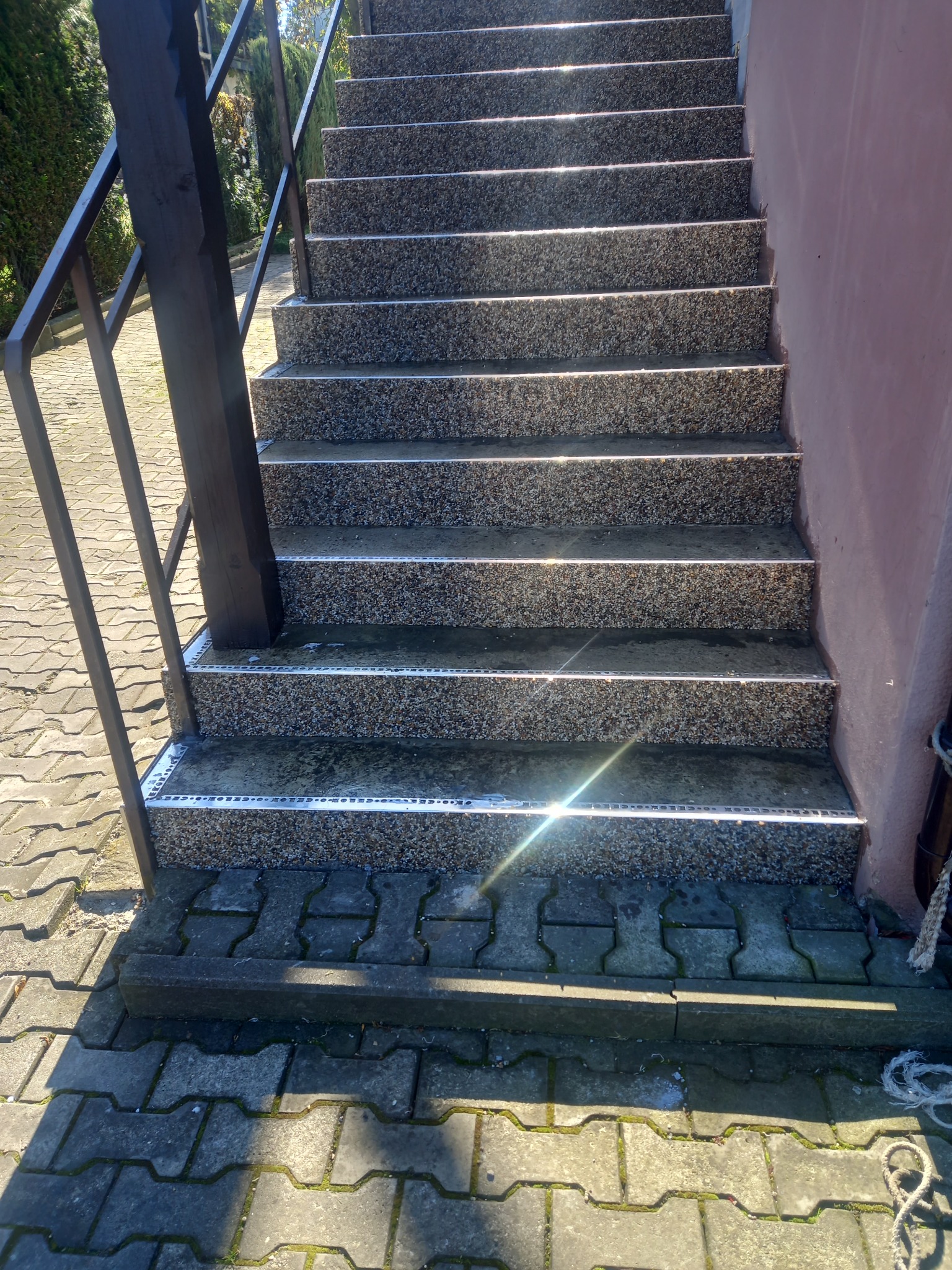 Zewnętrzne schody z szarego kamienia z aluminiowymi listwami zabezpieczającymi krawędzie, prowadzące do budynku o różowej elewacji. Widoczne metalowe poręcze i kostka brukowa na dole.