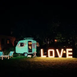 Turkusowa przyczepa z napisem 'PHOTO' na boku, oświetlona girlandami, stoi na trawie obok dużego, świecącego napisu 'LOVE', a w pobliżu znajdują się dwa leżaki z małym stolikiem, wszystko w nocnej...