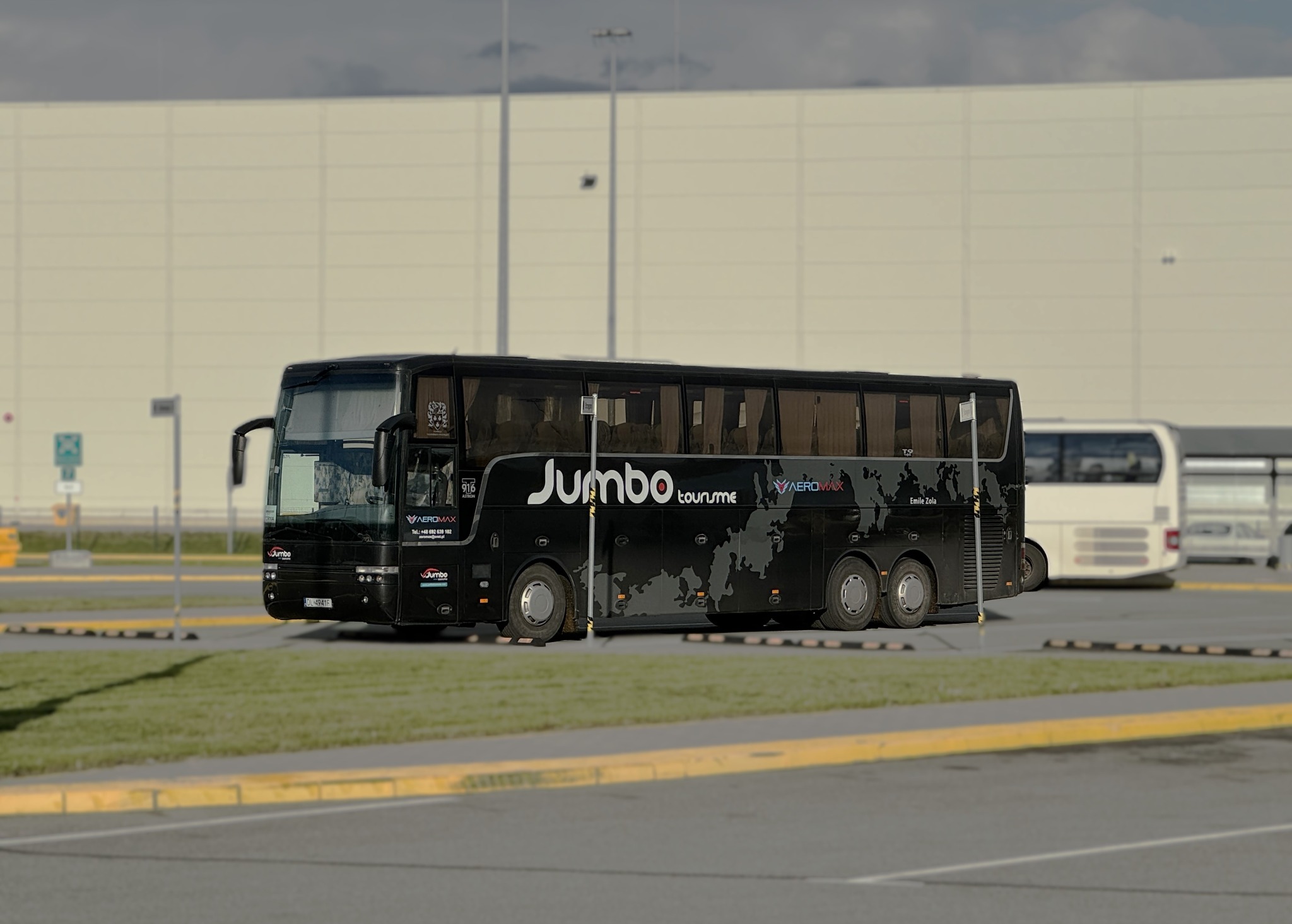 Czarny autokar turystyczny marki Jumbo Toursme z mapą świata na boku, zaparkowany na parkingu z żółtymi krawężnikami, na tle białego budynku magazynowego i innego, białego autobusu w oddali.