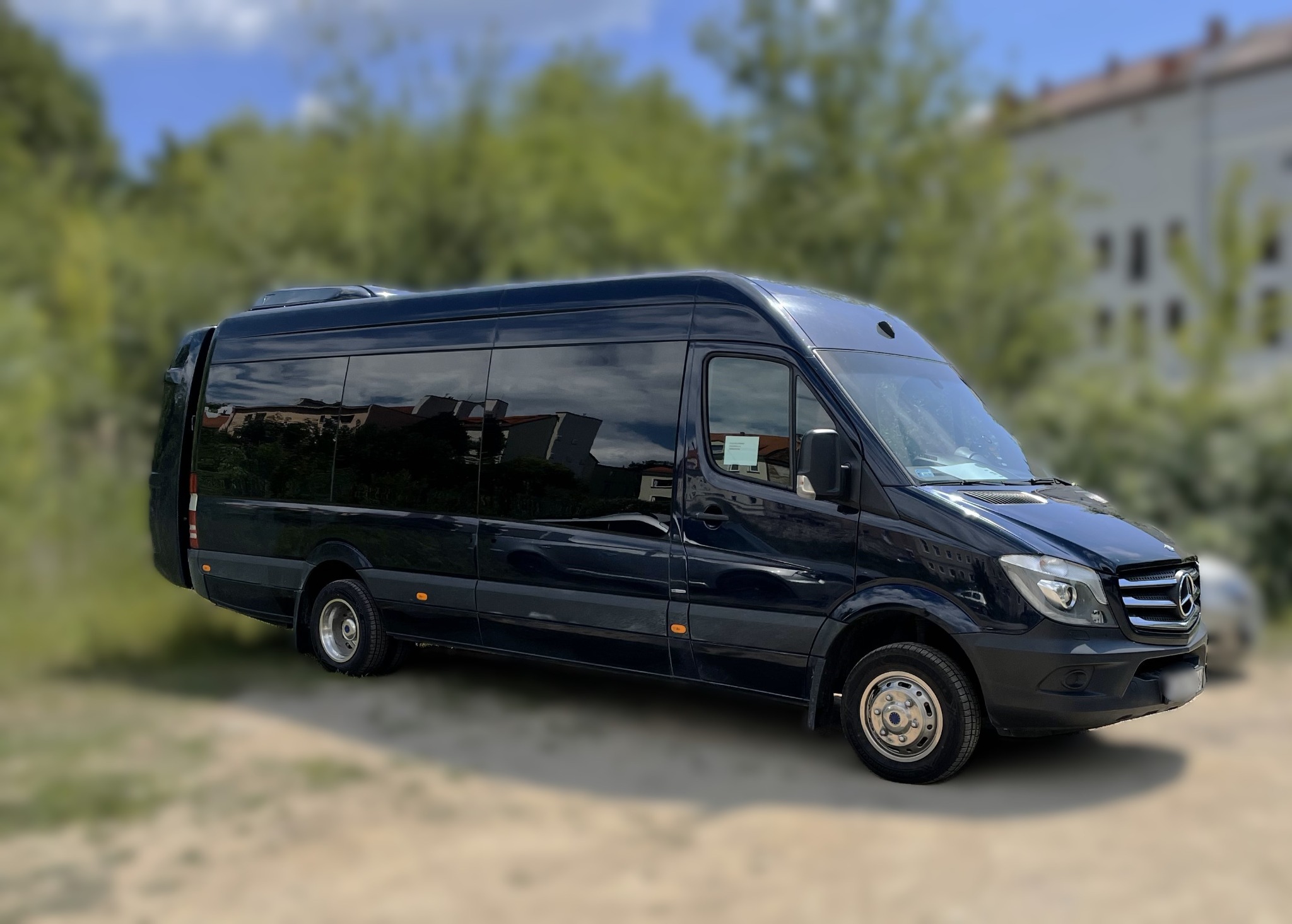 Czarny, błyszczący bus Mercedes-Benz Sprinter zaparkowany na żwirowym podłożu, z budynkiem odbijającym się w przyciemnianych szybach.