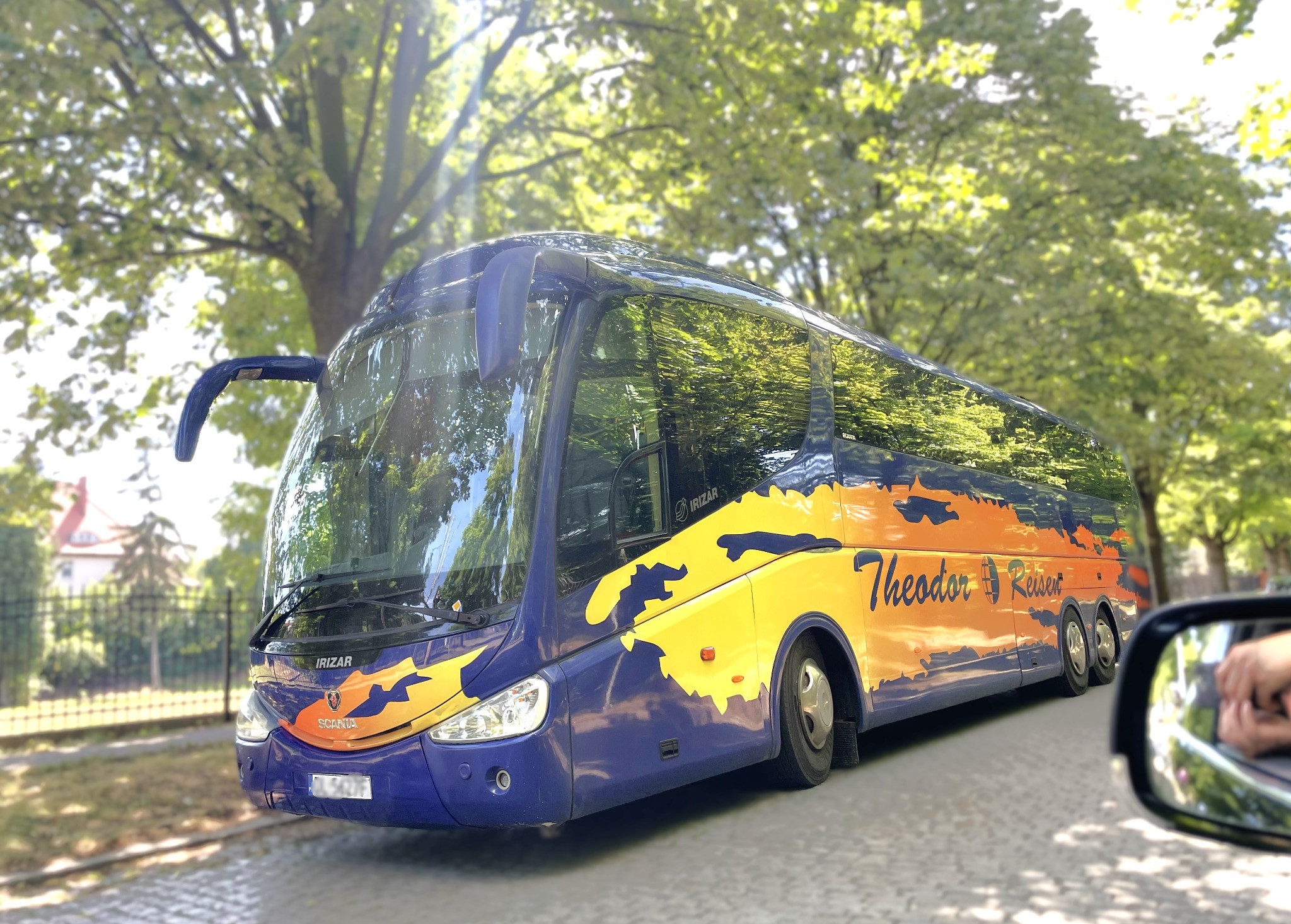 Niebiesko-żółty autokar turystyczny marki Irizar, z grafiką i nazwą 'Theodor Reisen', sfotografowany z perspektywy kierowcy samochodu, z widocznym lusterkiem bocznym i zielonym tłem drzew.