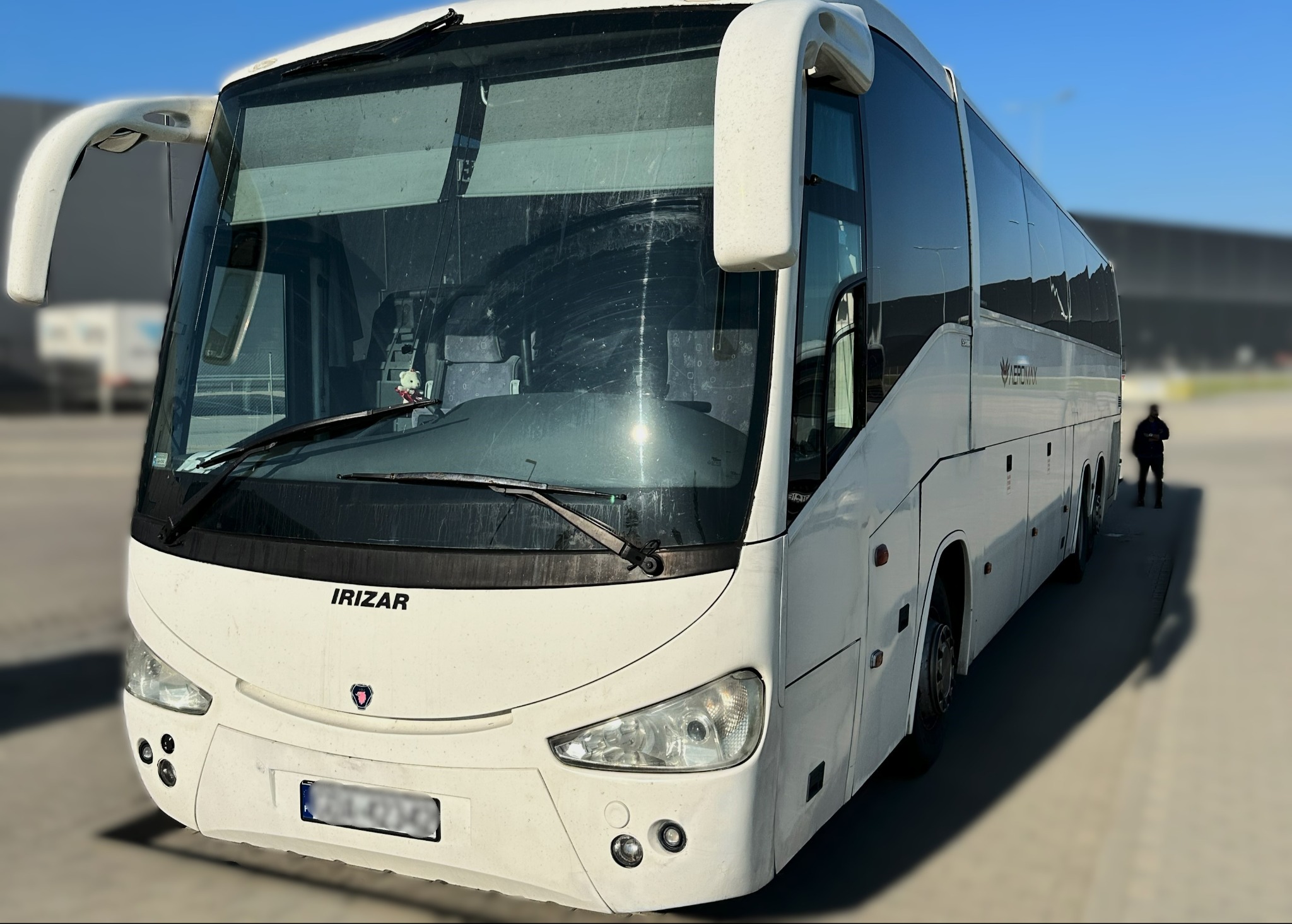 Biały autokar Irizar na parkingu, widok z przodu, z widoczną maskotką Hello Kitty na desce rozdzielczej oraz osobą w tle.