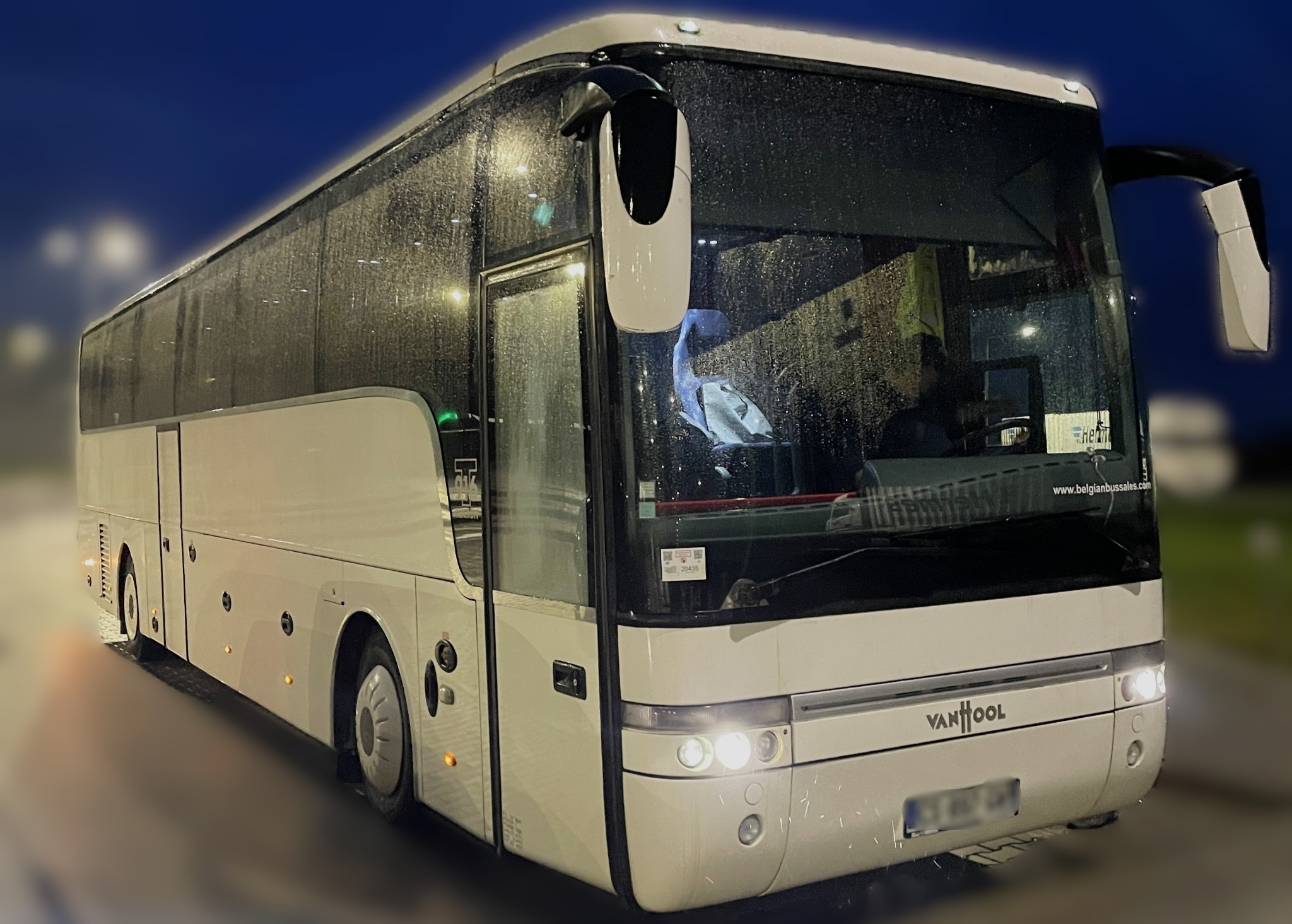 Biały autokar Van Hool z mokrą szybą przednią, widoczny kierowca w środku, nocne oświetlenie.