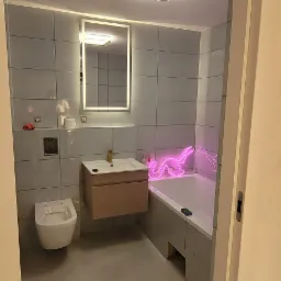 Wykończona łazienka z szarymi płytkami, podwieszaną toaletą, umywalką z szafką w kolorze drewna, podświetlanym lustrem i neonowym napisem nad wanną.