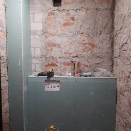 OL-MAL - Montaż stelaża podtynkowego WC z płyt karton-gips na tle surowej, częściowo otynkowanej ściany z cegły, widoczne narzędzia i elementy instalacji hydraulicznej.