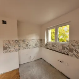 Wnętrze kuchni w trakcie remontu: ułożone płytki podłogowe w odcieniach szarości, częściowo położona dekoracyjna glazura na ścianach, widoczne instalacje elektryczne i hydrauliczne, fragment starej...