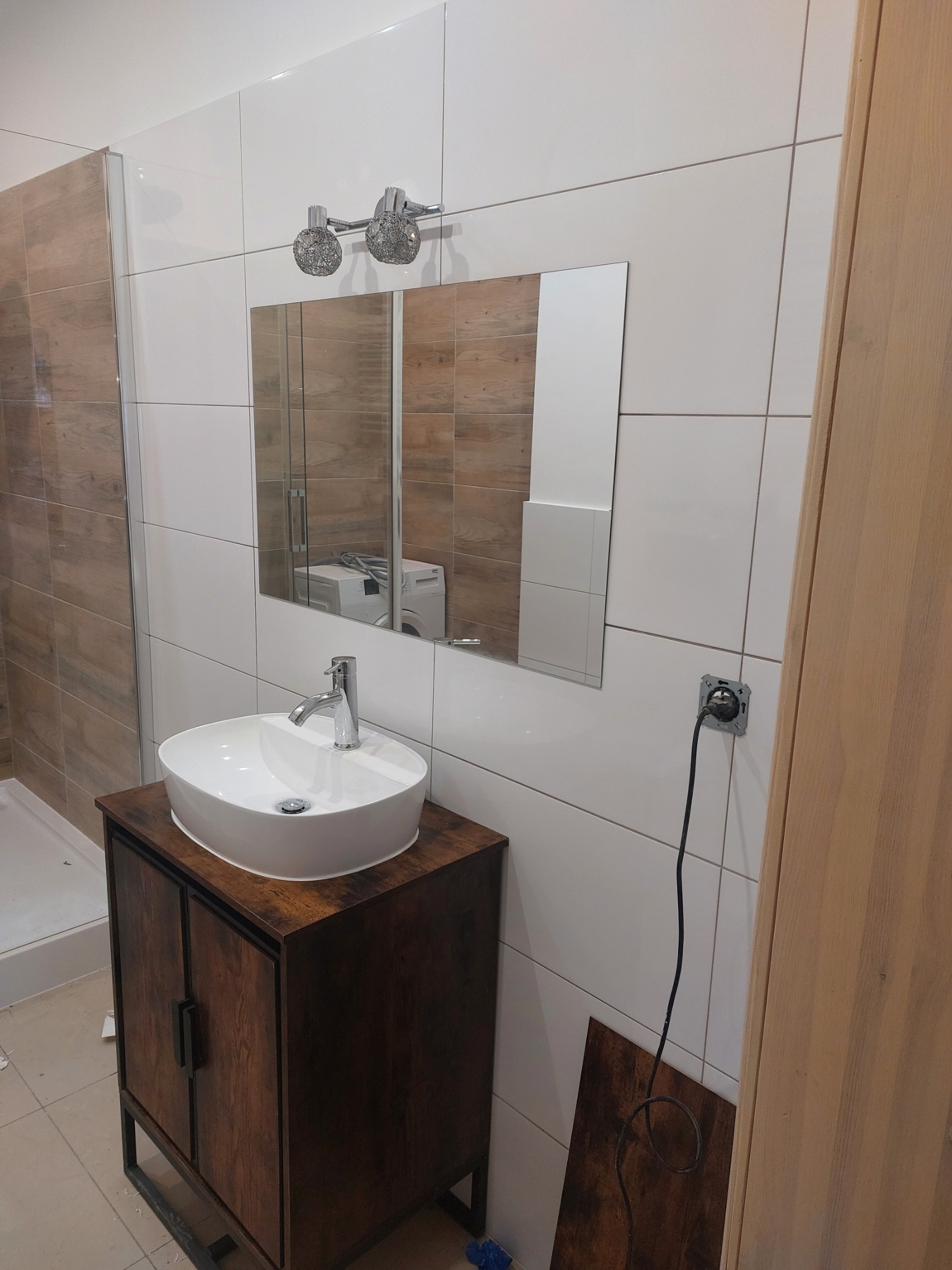 Nowoczesna łazienka z biało-brązowymi płytkami, owalna umywalka nablatowa na szafce w stylu rustykalnym, chromowana bateria, lustro odbijające pralkę, oraz ozdobne lampy nad lustrem. Widoczna...