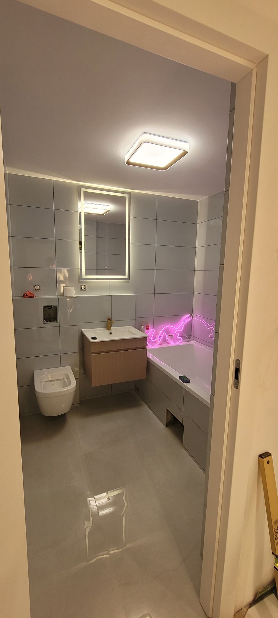 Wykończona łazienka z szarymi płytkami, podwieszaną toaletą, umywalką z szafką w kolorze drewna, podświetlanym lustrem i neonowym napisem nad wanną.