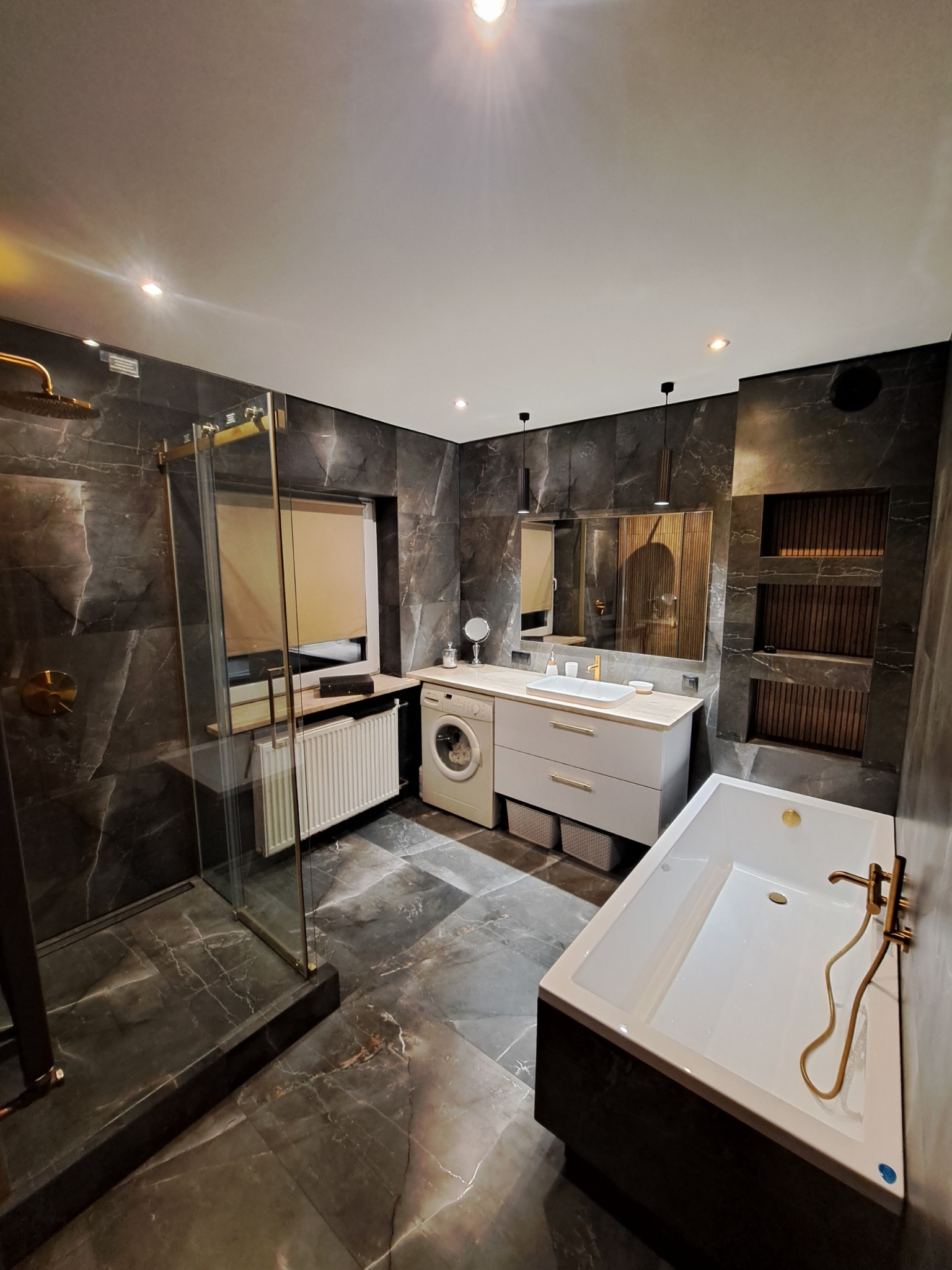 Nowoczesna łazienka z marmurowymi płytkami, kabiną prysznicową typu walk-in z armaturą w kolorze mosiądzu, białą wanną, umywalką nablatową, pralką i grzejnikiem żeberkowym.