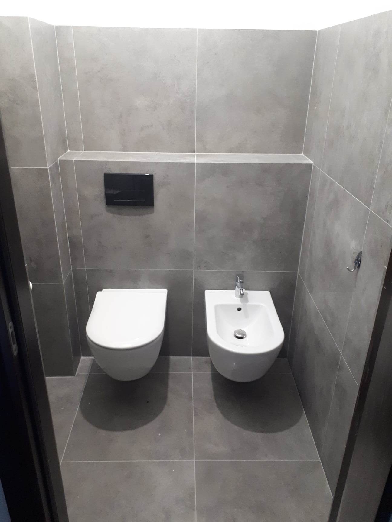 Wykończona toaleta z podwieszaną muszlą klozetową i bidetem w szarej, minimalistycznej aranżacji z płytek imitujących beton.