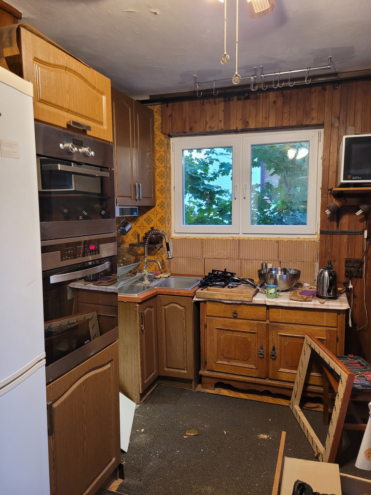 Kuchnia przed remontem: widoczne stare, drewniane szafki, przestarzałe urządzenia AGD, okno z widokiem na zieleń, ściany wyłożone boazerią i częściowo pomalowane na żółto, nieporządek na blacie...