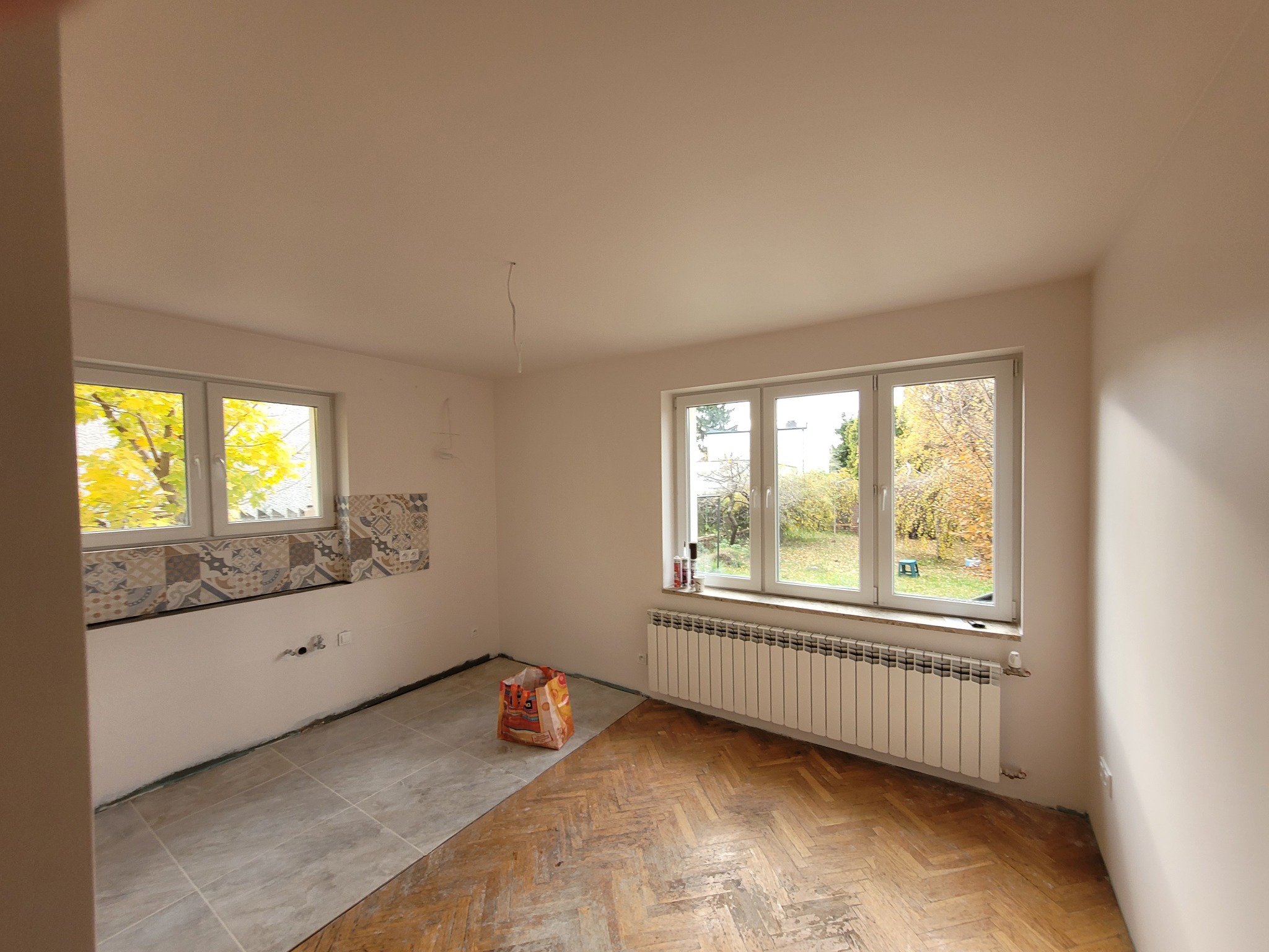 Pokój w trakcie remontu: częściowo ułożona szara terakota na podłodze, fragment boazerii z płytek pod oknem, odsłonięta stara klepka parkietowa, grzejnik pod oknem, widoczne przewody elektryczne.