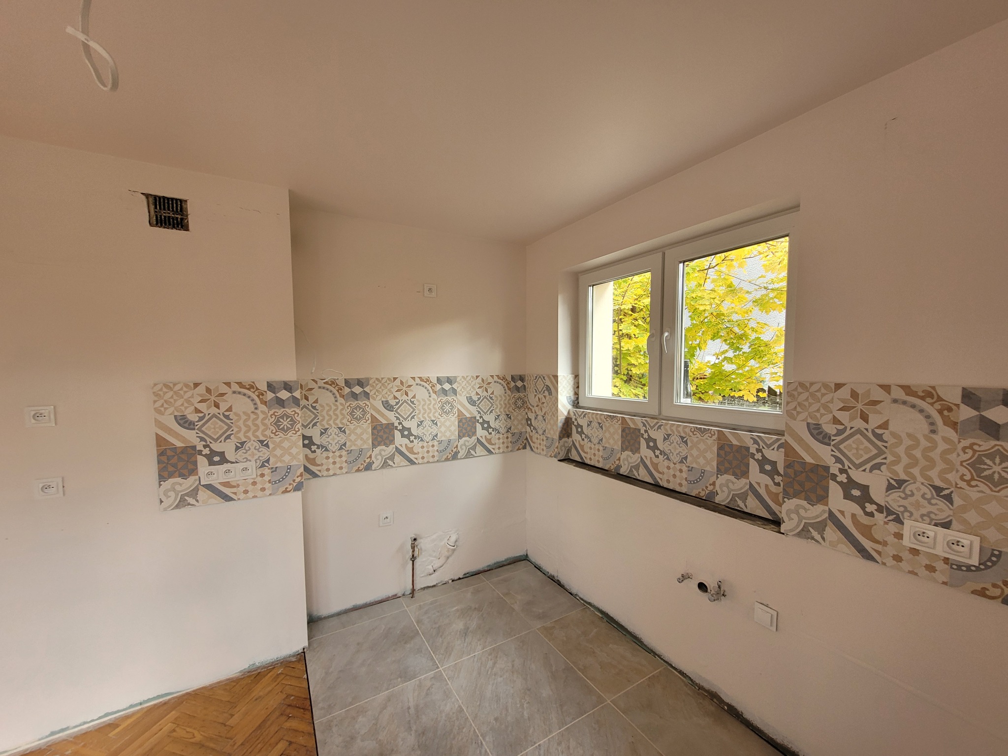 Wnętrze kuchni w trakcie remontu: ułożone płytki podłogowe w odcieniach szarości, częściowo położona dekoracyjna glazura na ścianach, widoczne instalacje elektryczne i hydrauliczne, fragment starej...