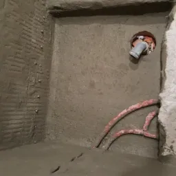 Surowy beton pokrywający ściany i podłogę w pomieszczeniu, widoczne rury i otw&oacute;r z wystającą rurą, ślady po poziomowaniu na ścianie, metalowe kołki w ścianie.