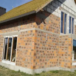 Budowa domu jednorodzinnego w trakcie montażu okien, widoczna surowa elewacja z cegły, częściowo ocieplony dach i drabina oparta o ścianę.