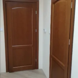 Dwie brązowe, wewnętrzne drzwi z płycinami w jasnym korytarzu z panelami podłogowymi imitującymi drewno, widoczne klamki i zawiasy.