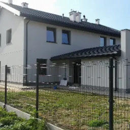 Nowoczesny dom jednorodzinny z szarymi elewacjami, czarną dachówką i ogrodzeniem panelowym, widok z zewnątrz w słoneczny dzień.