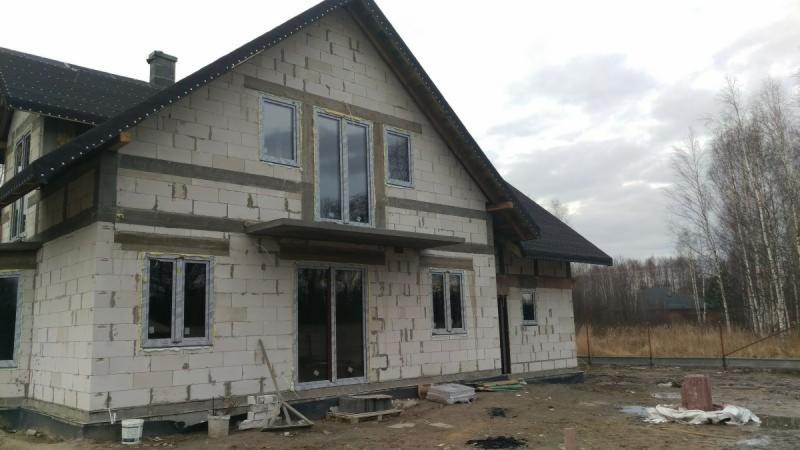 Surowy stan budynku jednorodzinnego z zamontowanymi oknami i drzwiami przesuwnymi, widoczne bloczki betonowe i świeże obramowania okienne, w tle las brzozowy.