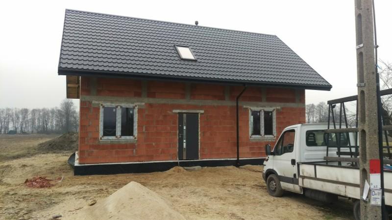 Surowy stan budynku z cegły z zamontowanymi oknami i drzwiami, widoczny samochód dostawczy na tle piaszczystego terenu.