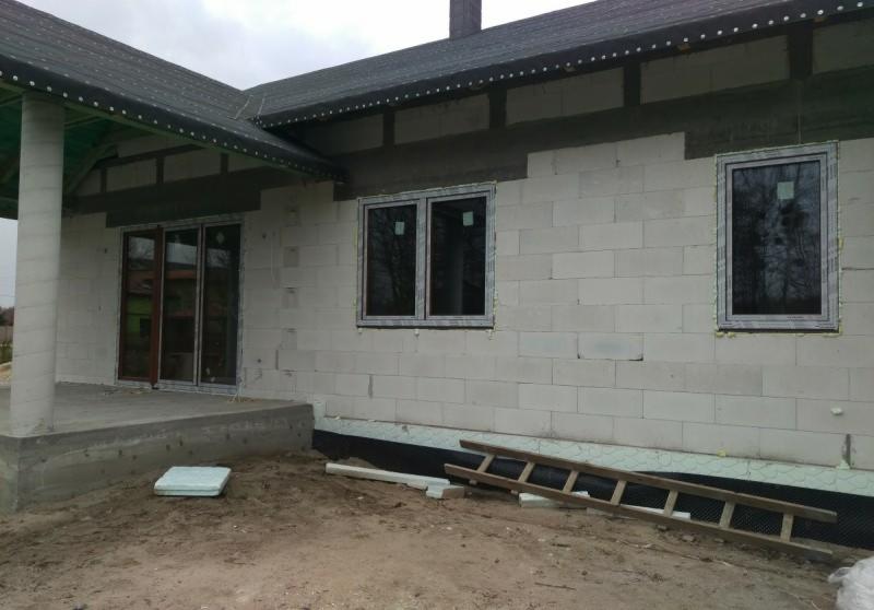 Dom w trakcie budowy: widoczne zamontowane okna, drzwi balkonowe, ściany z bloczków betonowych, przygotowane do ocieplenia, oraz drewniana drabina oparta o fundament.