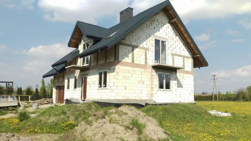 Dom w trakcie budowy z białego pustaka, z zamontowanymi oknami i drzwiami, ciemny dach z blachy, widok z zewnątrz na tle łąki i błękitnego nieba z chmurami.