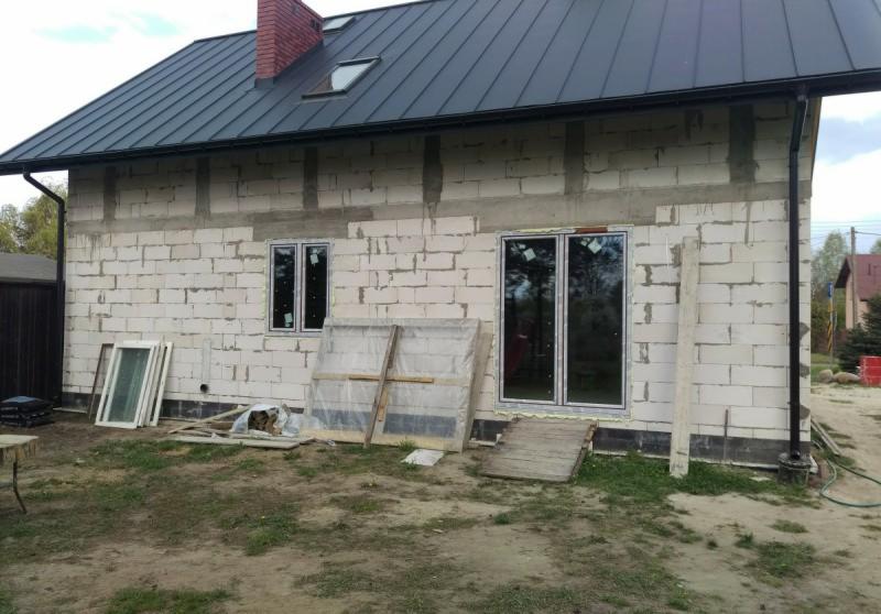 Dom w trakcie budowy z zamontowanymi oknami i drzwiami przesuwnymi, widoczne okna zabezpieczone folią, na ziemi leżą pozostałości po montażu.