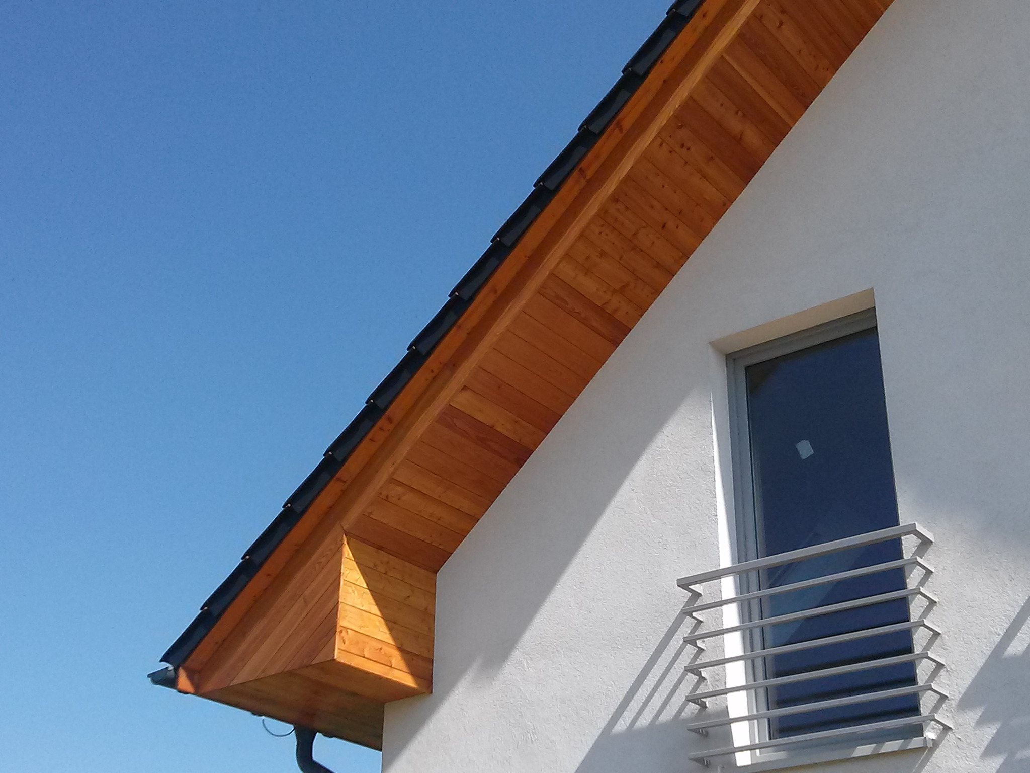 Fragment nowego dachu z drewnianą podbitką i czarną dachówką na tle błękitnego nieba, widok z dołu.