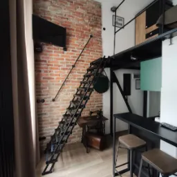 Loftowe mieszkanie z metalowymi schodami prowadzącymi na antresolę, ceglaną ścianą i starą maszyną do szycia Singer jako elementem dekoracyjnym.