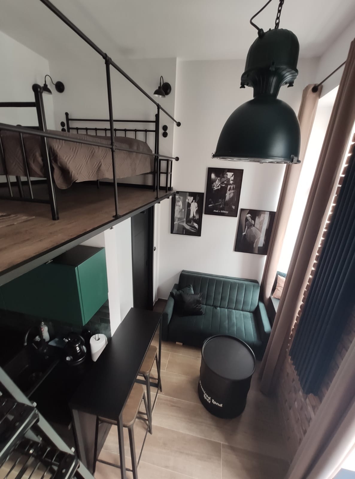 Loftowe mieszkanie z antresolą sypialną, metalowymi schodami, zieloną sofą, czarnym stolikiem-beczką i wiszącą lampą w stylu industrialnym.