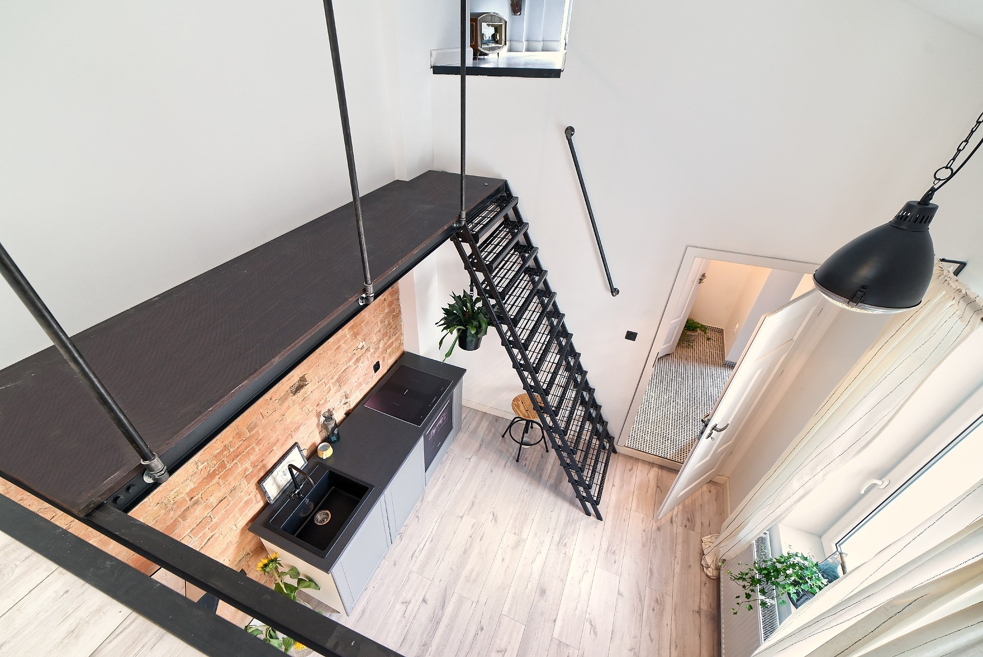 Loft z antresolą: czarne metalowe schody prowadzą na platformę z ceglaną ścianą i aneksem kuchennym, widok z góry na jasną podłogę i okno z zasłonami.