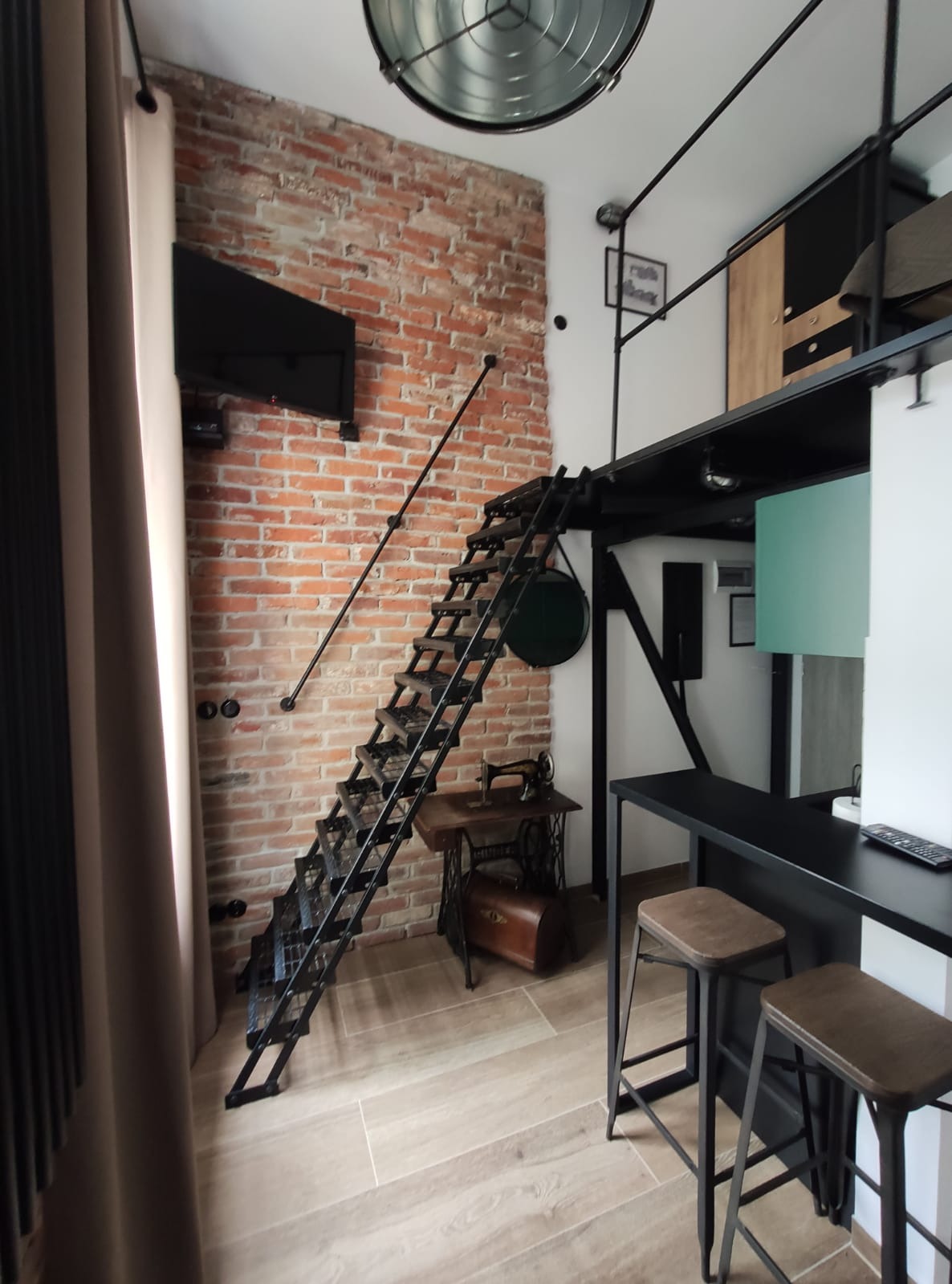Loftowe mieszkanie z metalowymi schodami prowadzącymi na antresolę, ceglaną ścianą i starą maszyną do szycia Singer jako elementem dekoracyjnym.